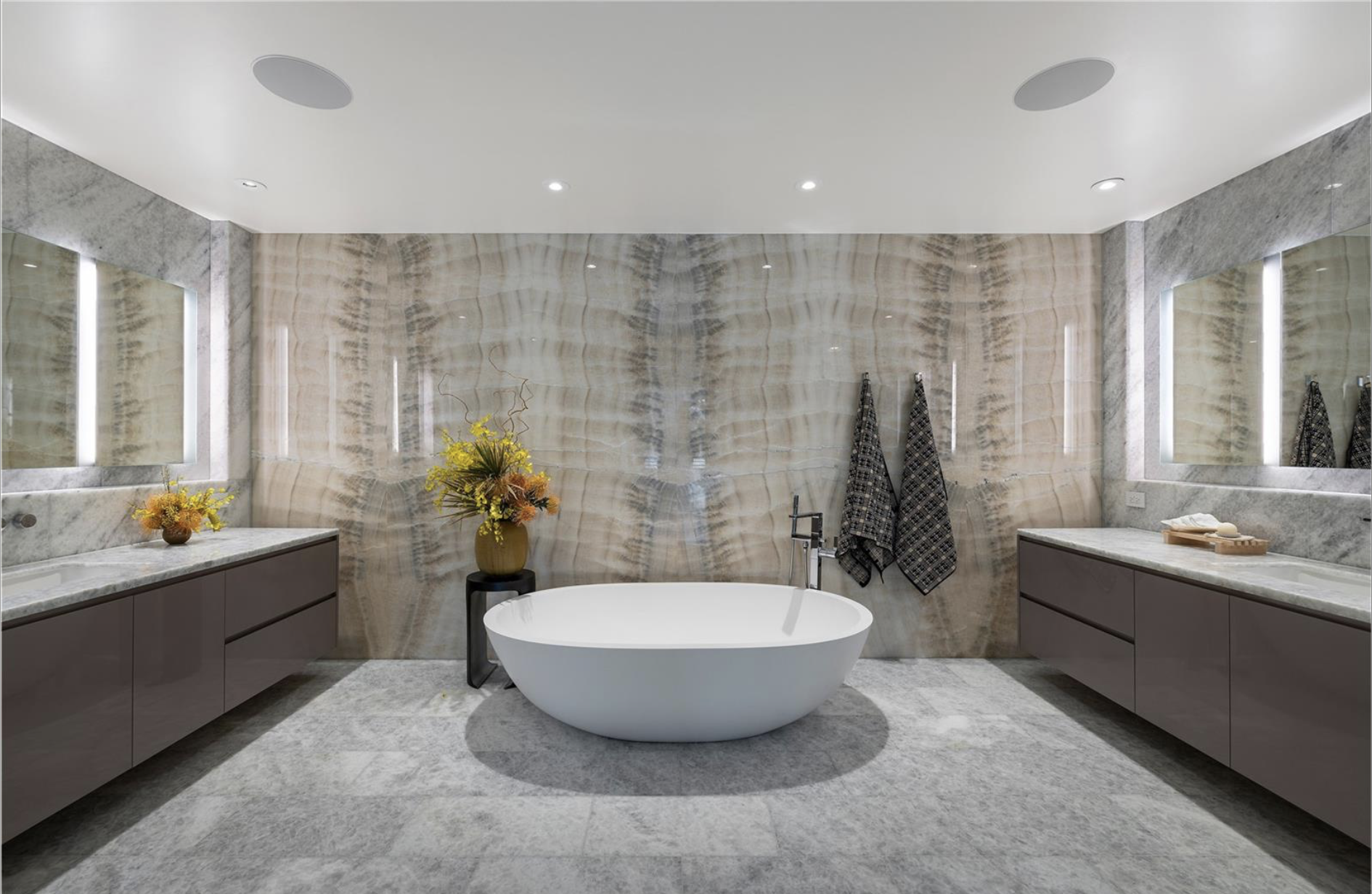 A modern bathroom with a freestanding bathtub centered on a large marble floor, flanked by grey vanities with marble countertops. The back wall features a polished stone design, with two towels hanging on the right side, and decorative flowers on the left.