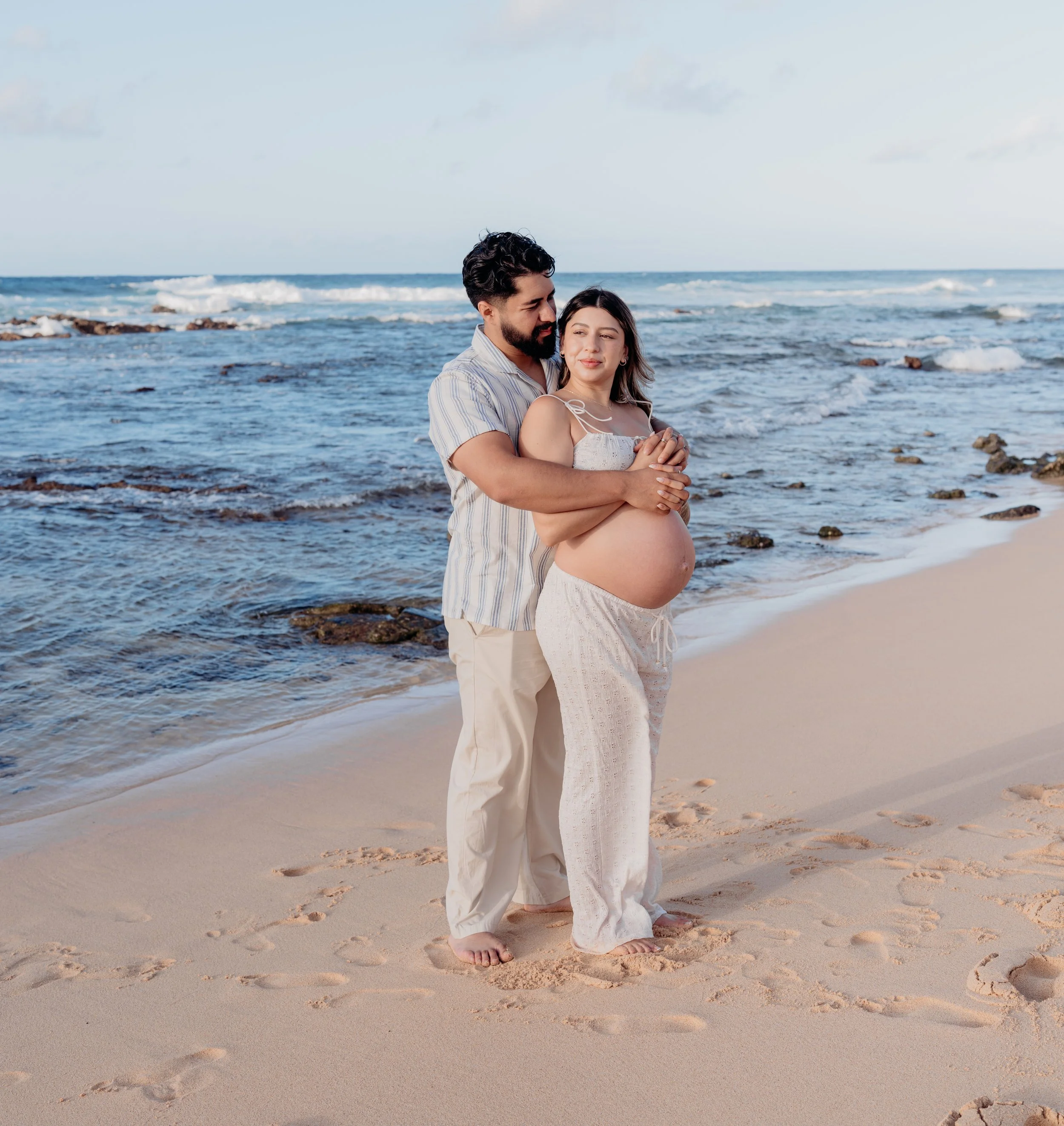 A pregnant woman and a man standing on the beach facing the ocean. The man is embracing the woman from behind, placing his hands on her belly. The woman is wearing white, and the man is wearing a short-sleeved, striped shirt and light-colored pants. 
