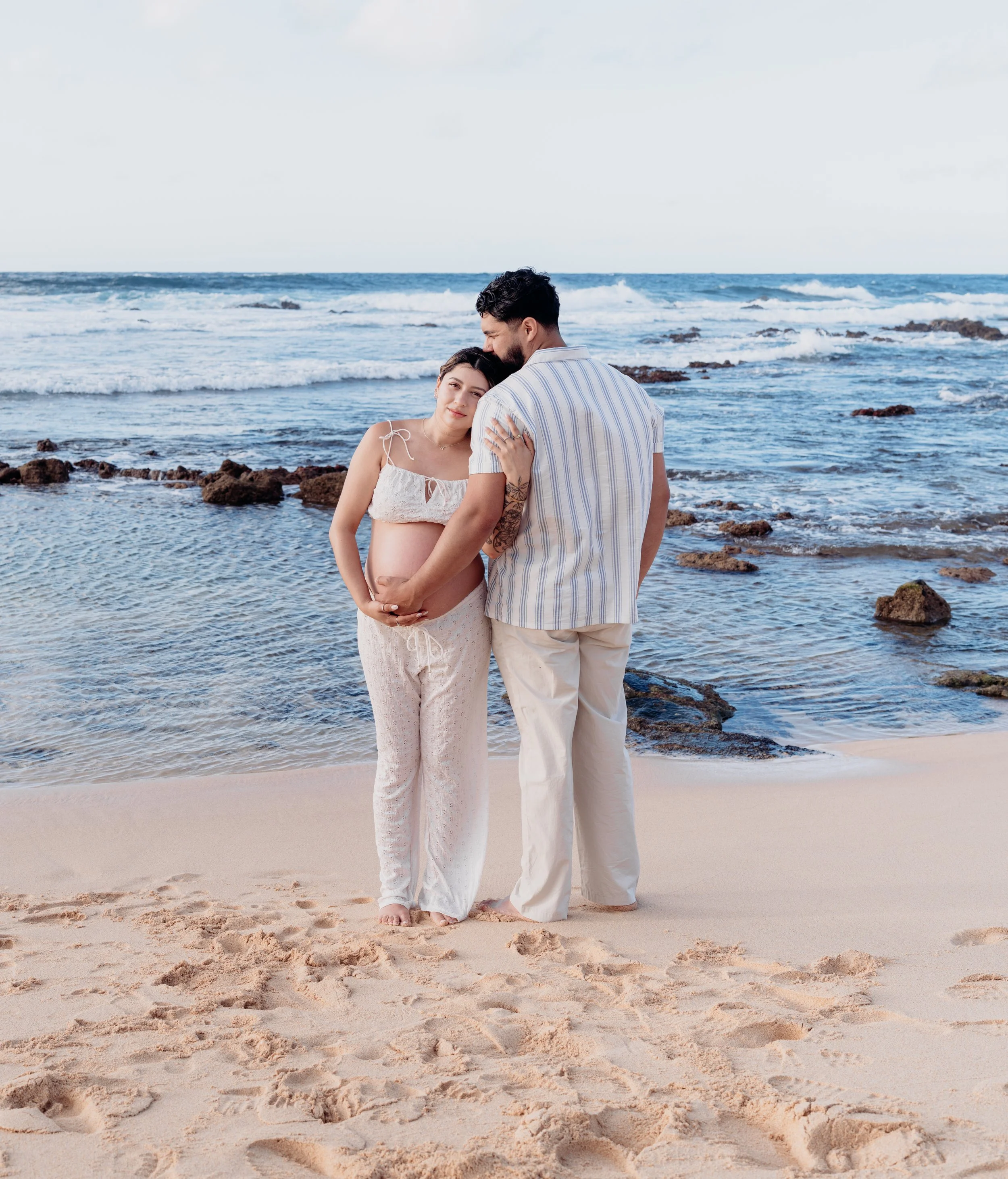 A pregnant woman and a man standing close on a sandy beach with ocean waves in the background. She is embracing her belly and they are holding hands, the woman leaning her head on his shoulder.