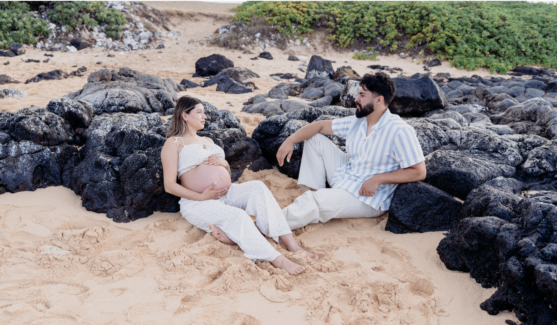 A pregnant woman and a man sitting on the beach among rocks, facing each other with relaxed expressions.