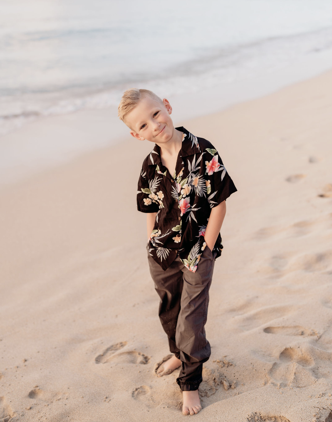 A young boy with blond hair smiling and walking on a sandy beach near the ocean, wearing a black Hawaiian shirt with floral patterns and dark pants.