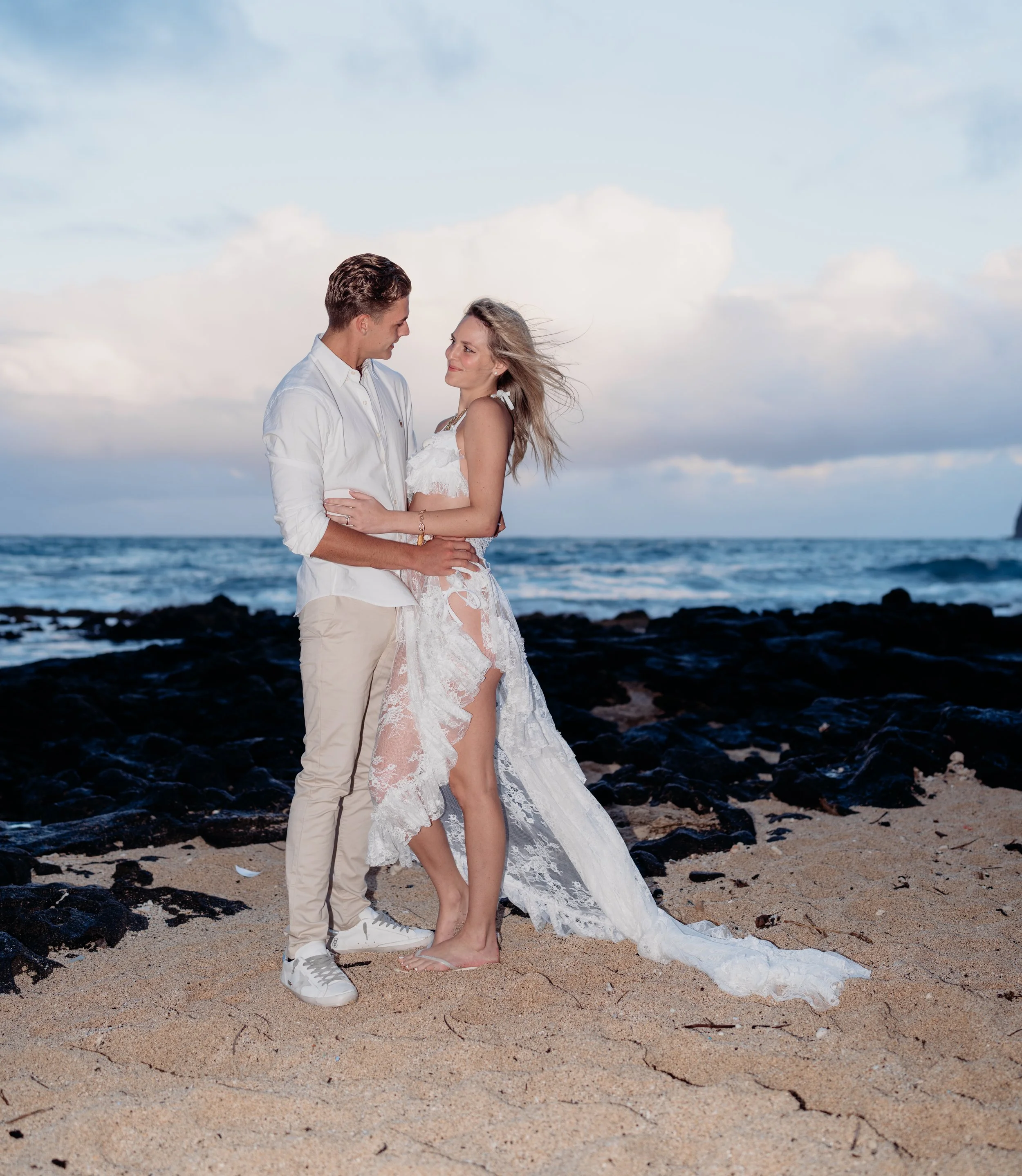 A couple on a beach, facing each other and smiling, with the ocean and a cloudy sky in the background. The woman is in a white lace dress, and the man is in a white shirt and beige pants.
