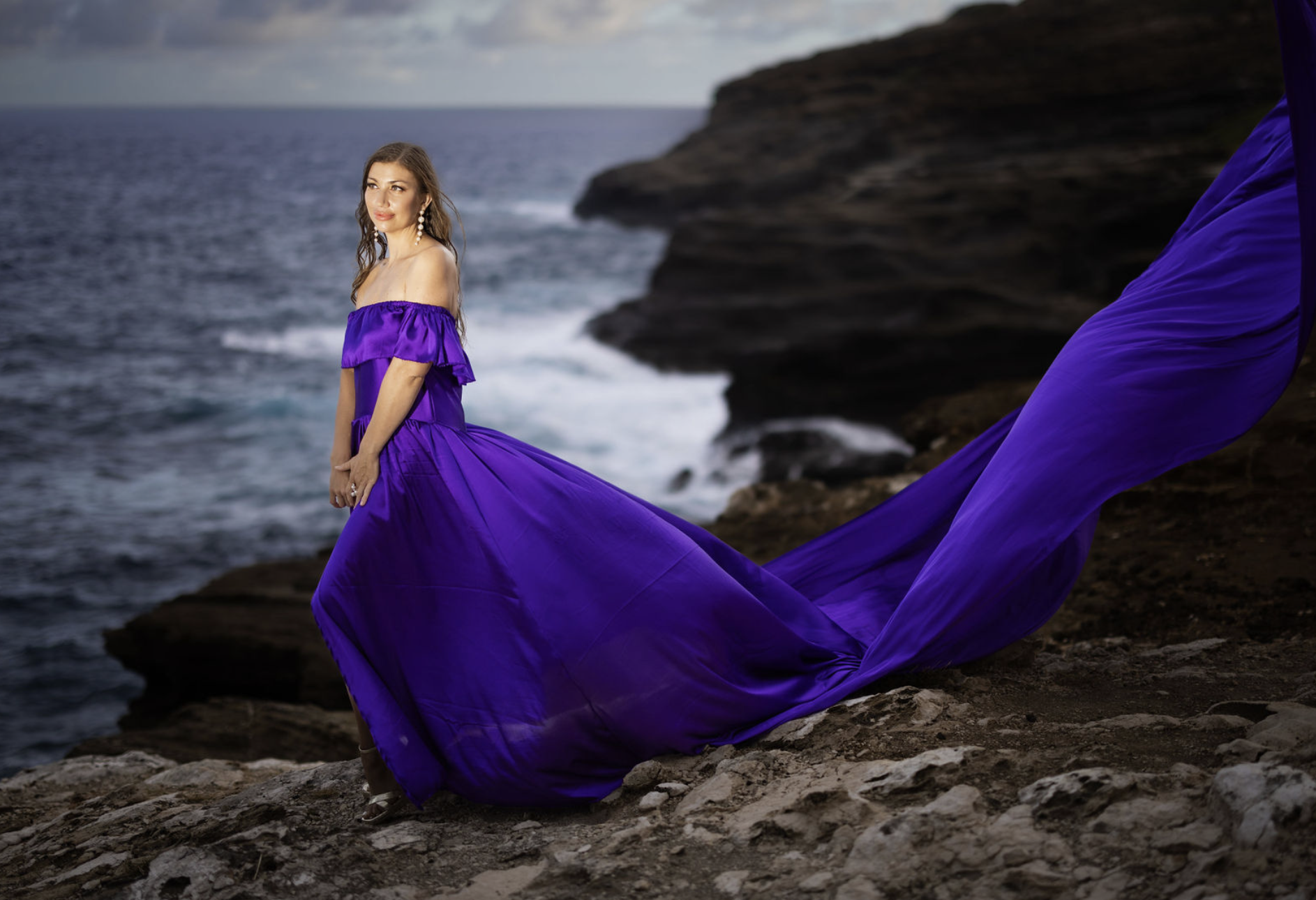 Flying dress photoshoot with a woman in a flowing purple gown on a dramatic ocean cliff
