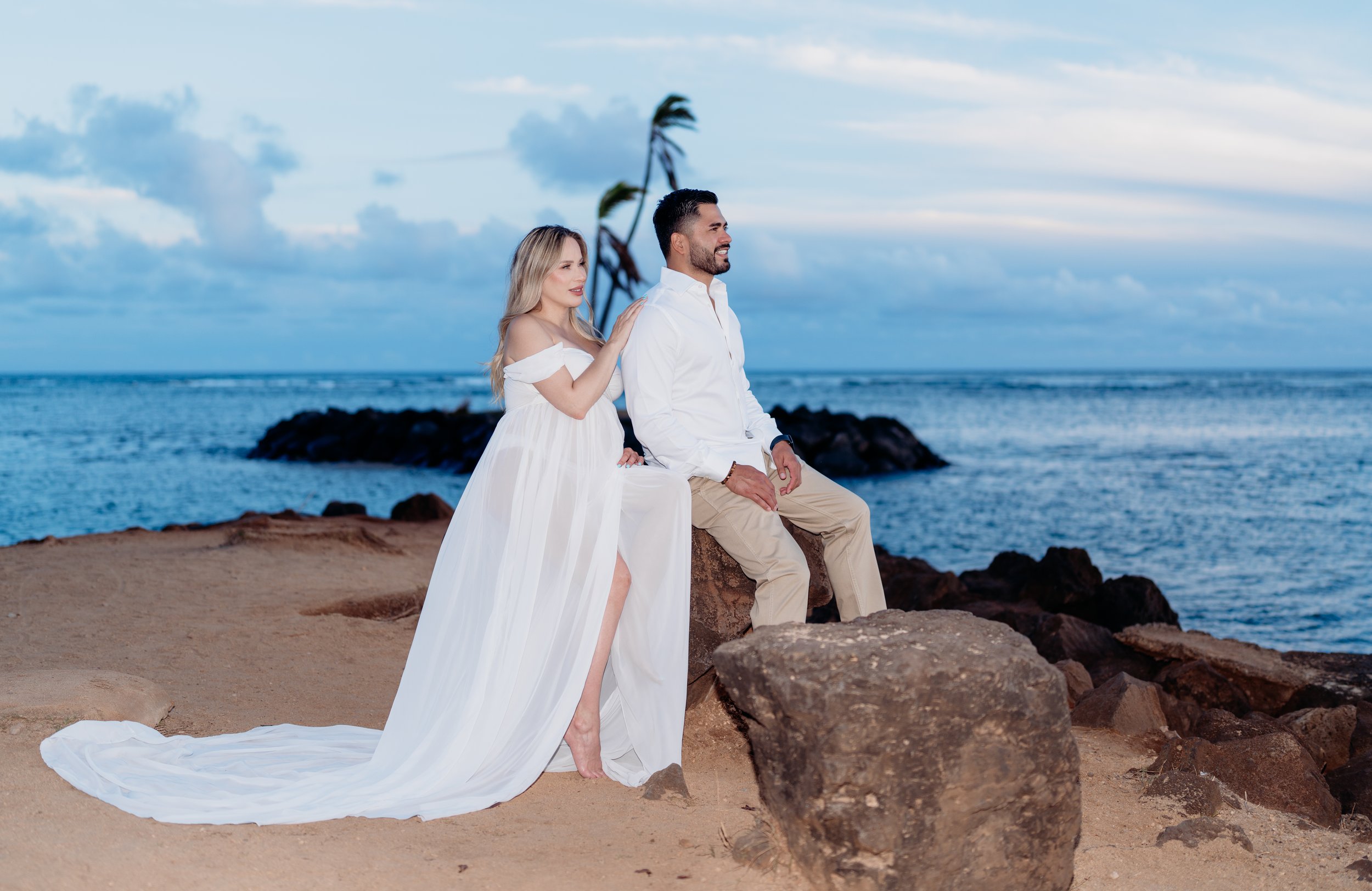 A couple dressed in white wearing wedding attire, posing on a beach with rocks and ocean in the background, under a cloudy sky.