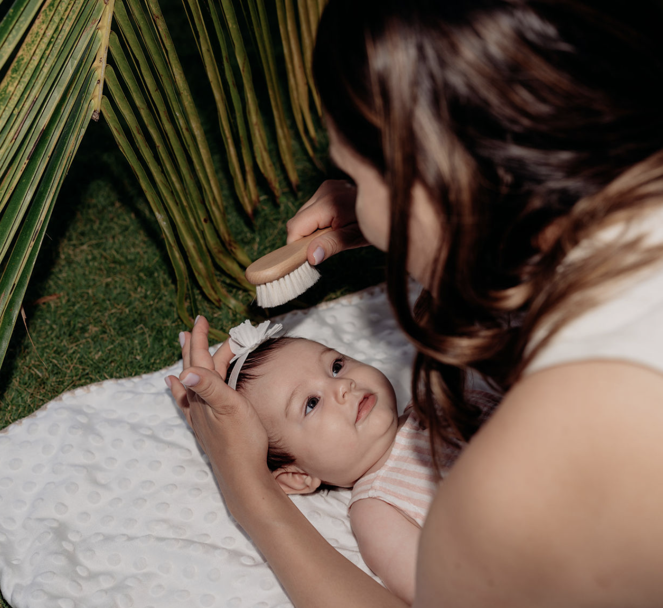 A woman grooming a baby with a brush outdoors, with green grass and palm leaves in the background.