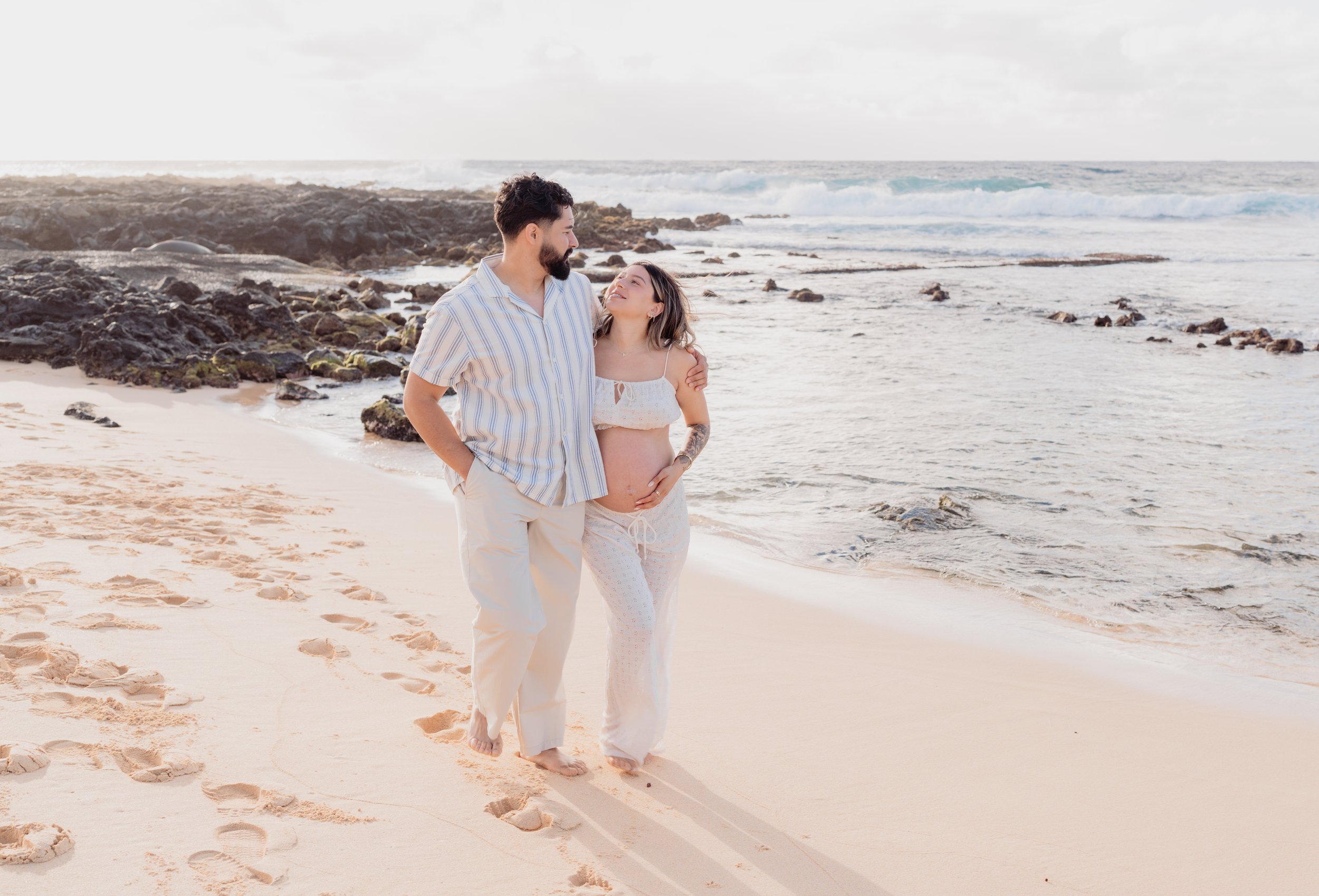 A pregnant woman and a man walking on a sandy beach during sunset, with rocks and waves in the background.