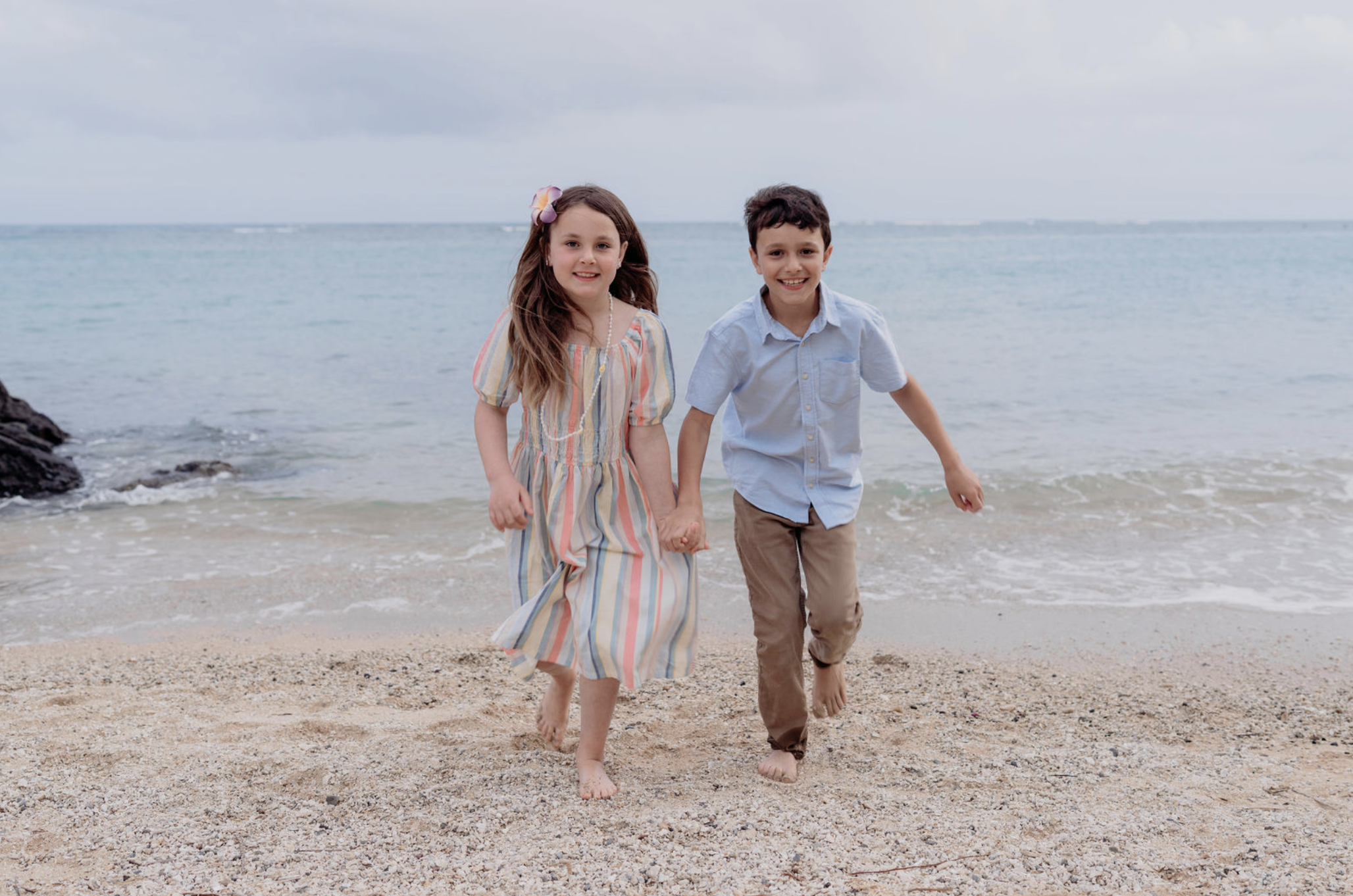 Two children, a girl and a boy, holding hands and running along the beach near the water with waves and rocks in the background.