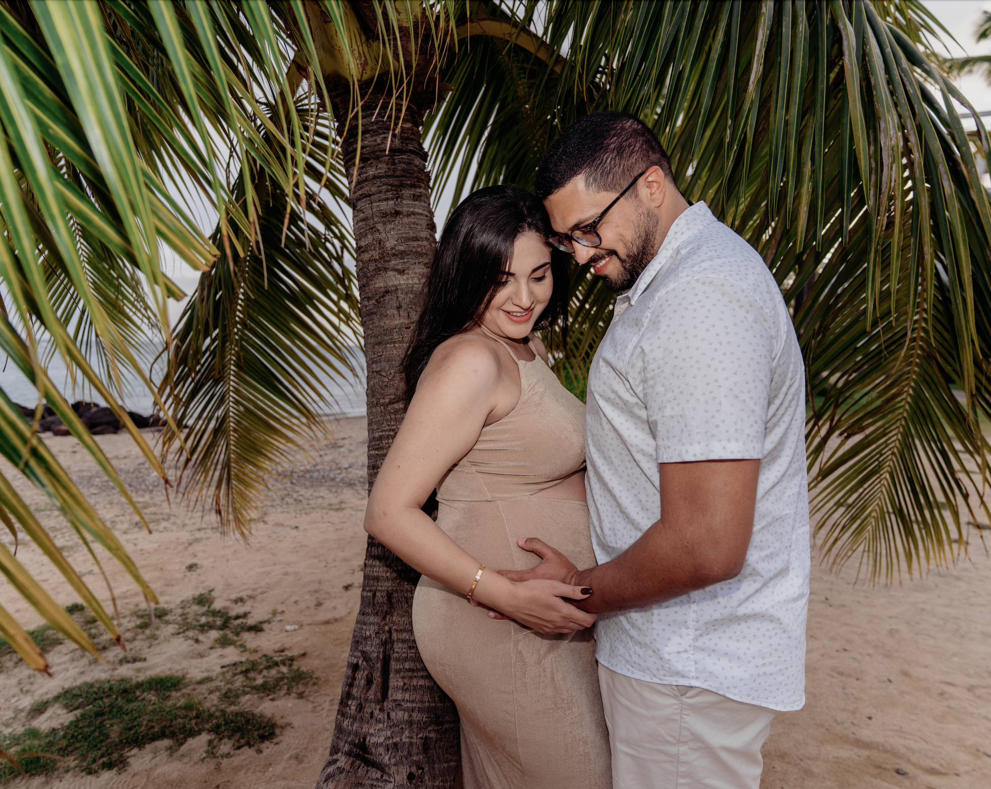 A pregnant woman and a man are sharing a tender moment under a palm tree on a sandy beach, smiling and holding her belly gently.