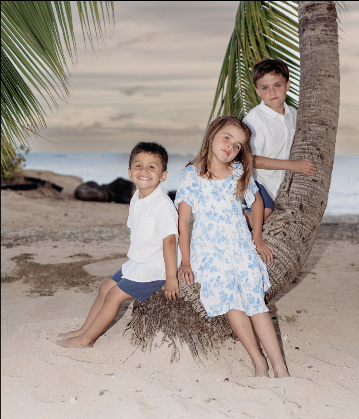 Three children on a beach with a palm tree and ocean in the background. One boy is sitting on the sand, smiling at the camera; a girl is sitting on the tree trunk with a relaxed expression; another boy is peeking out from behind the tree, holding ont