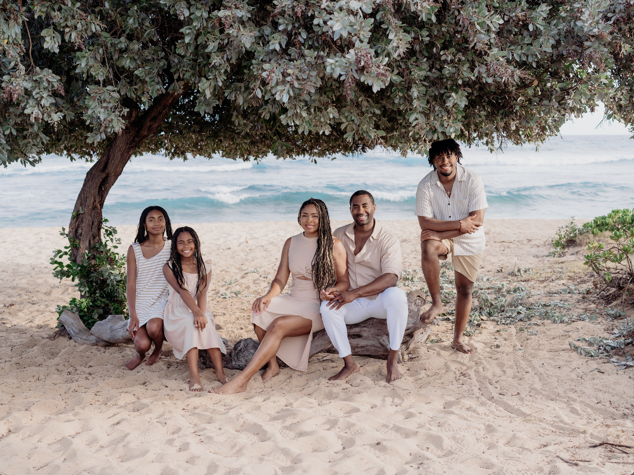 Family sitting and standing under a large tree on the beach, with the ocean in the background.
