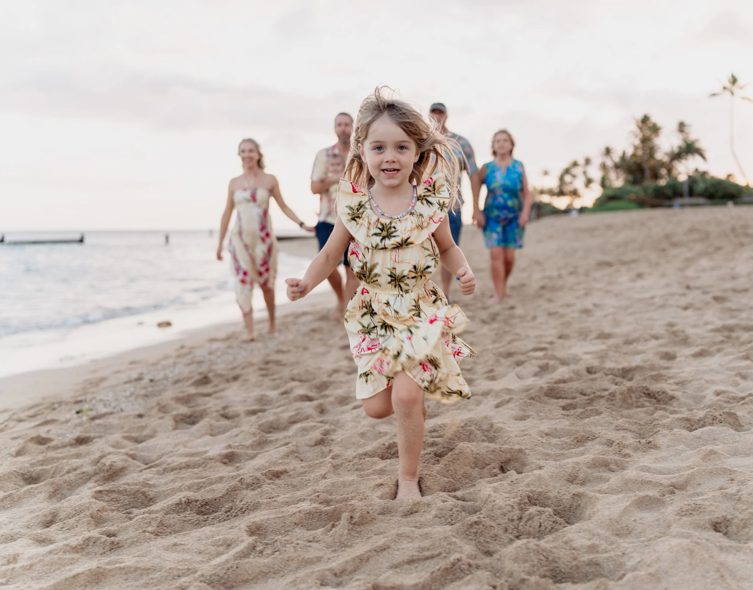A young girl in a floral dress runs towards the camera on a sandy beach, with a group of adults walking behind her near the shoreline and palm trees in the background.