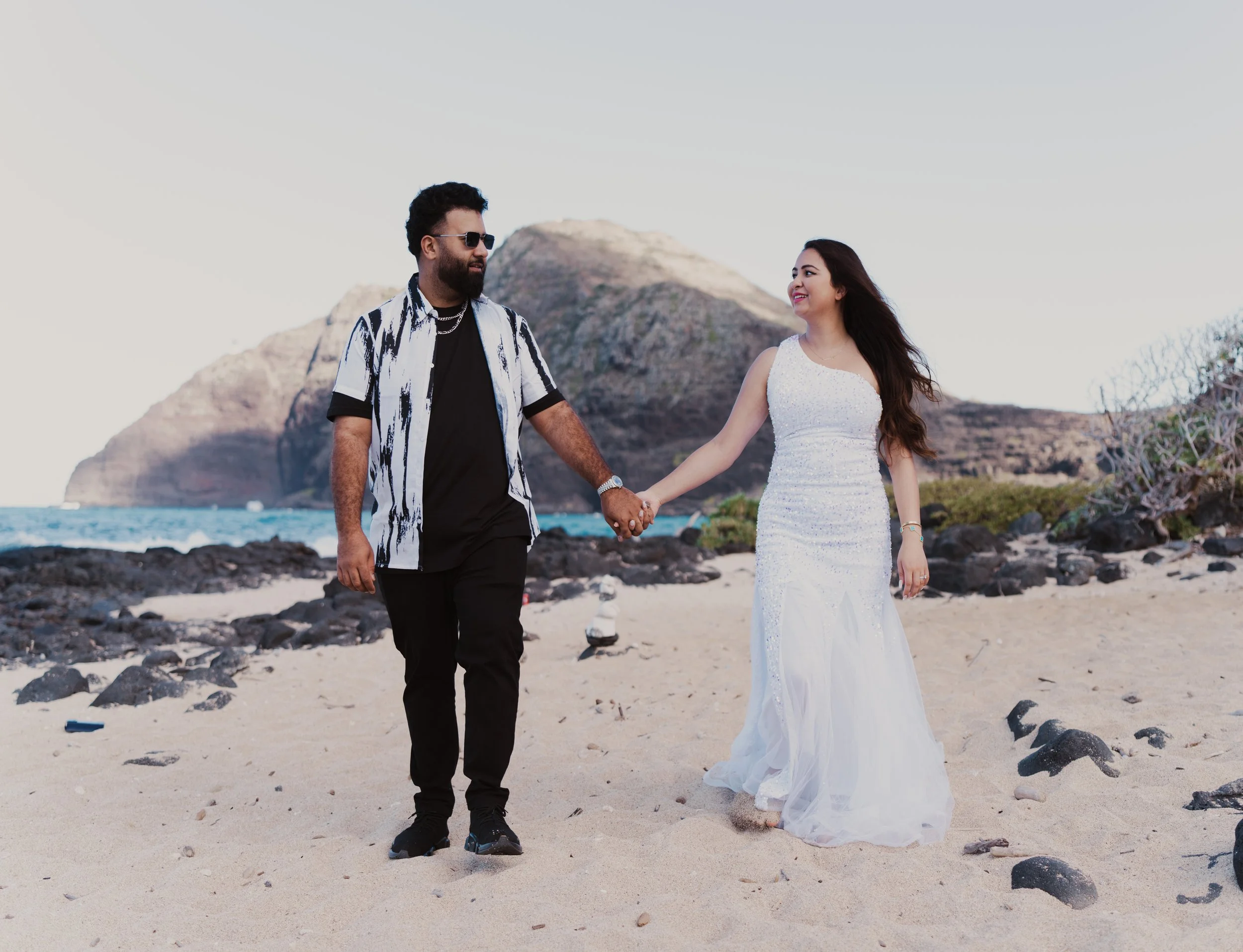 A couple holding hands on a beach, with mountains in the background. The woman is wearing a white dress and the man is wearing black pants and a patterned shirt.