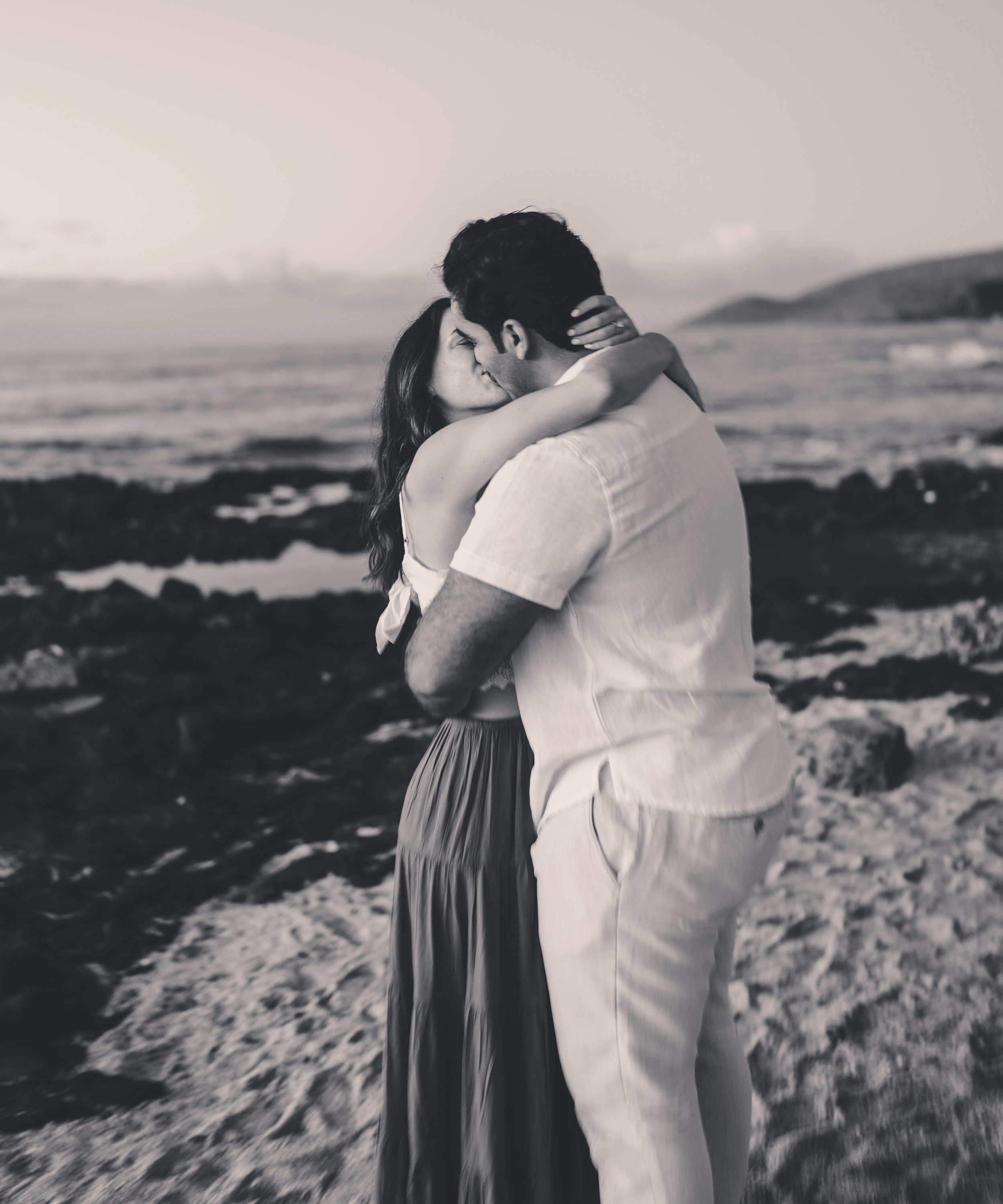 A black and white photo of a man and woman embracing and kissing on a beach at sunset.
