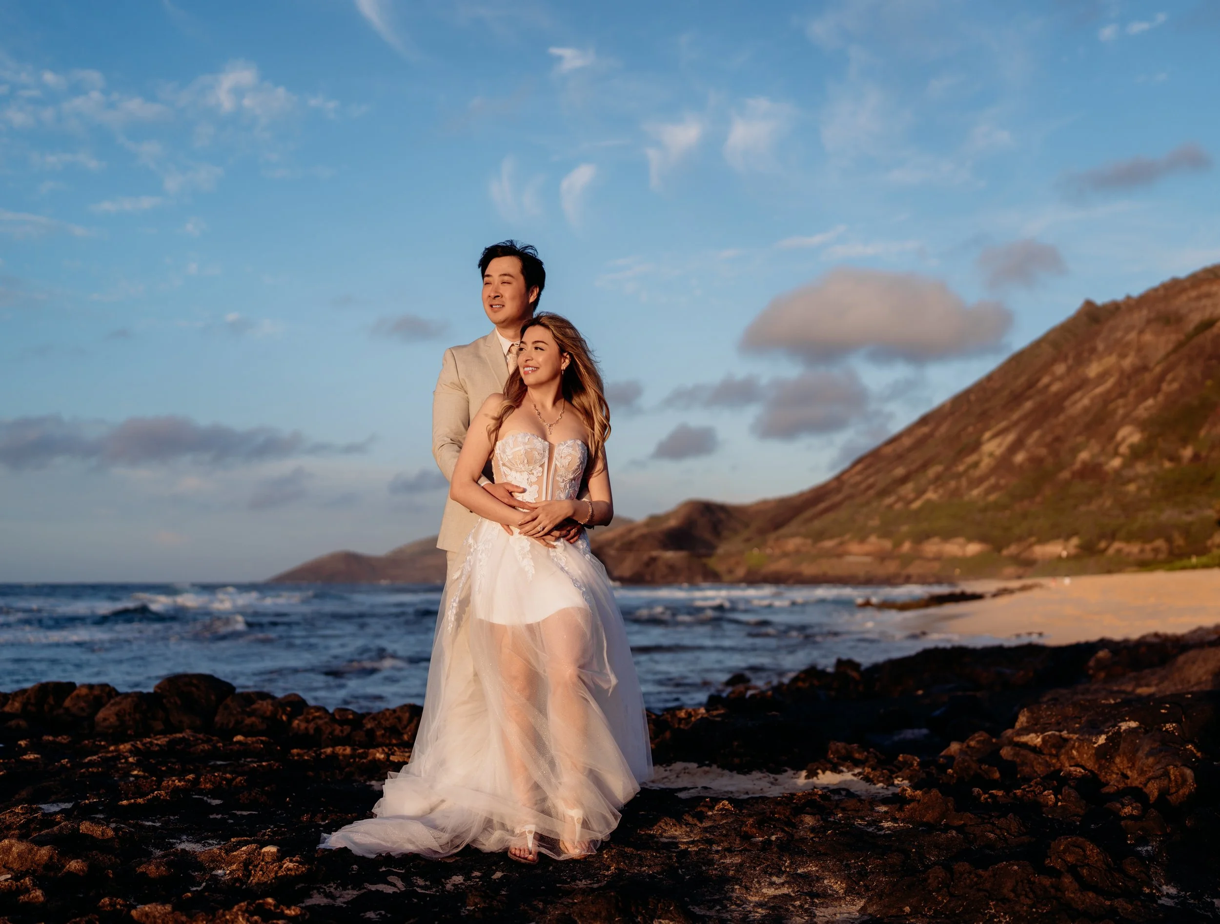 A couple in wedding attire standing on a rocky beach at sunset, with waves and a mountain in the background.