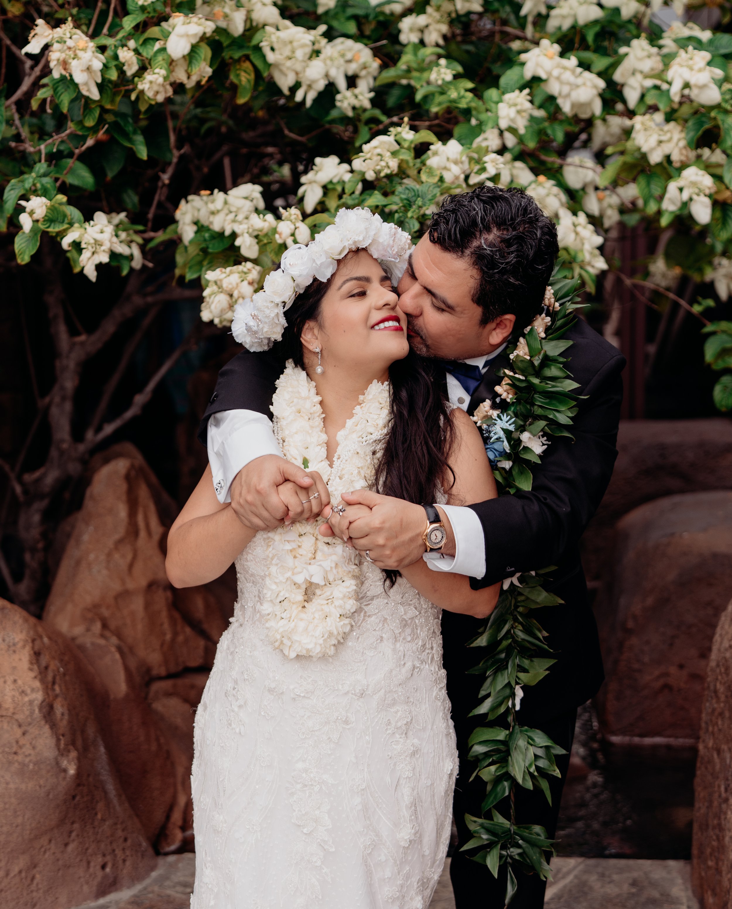 Couple kissing during their wedding, with the bride wearing a flower crown and lei, and the groom in a black tuxedo with a green lei, standing in front of white flowering bushes.