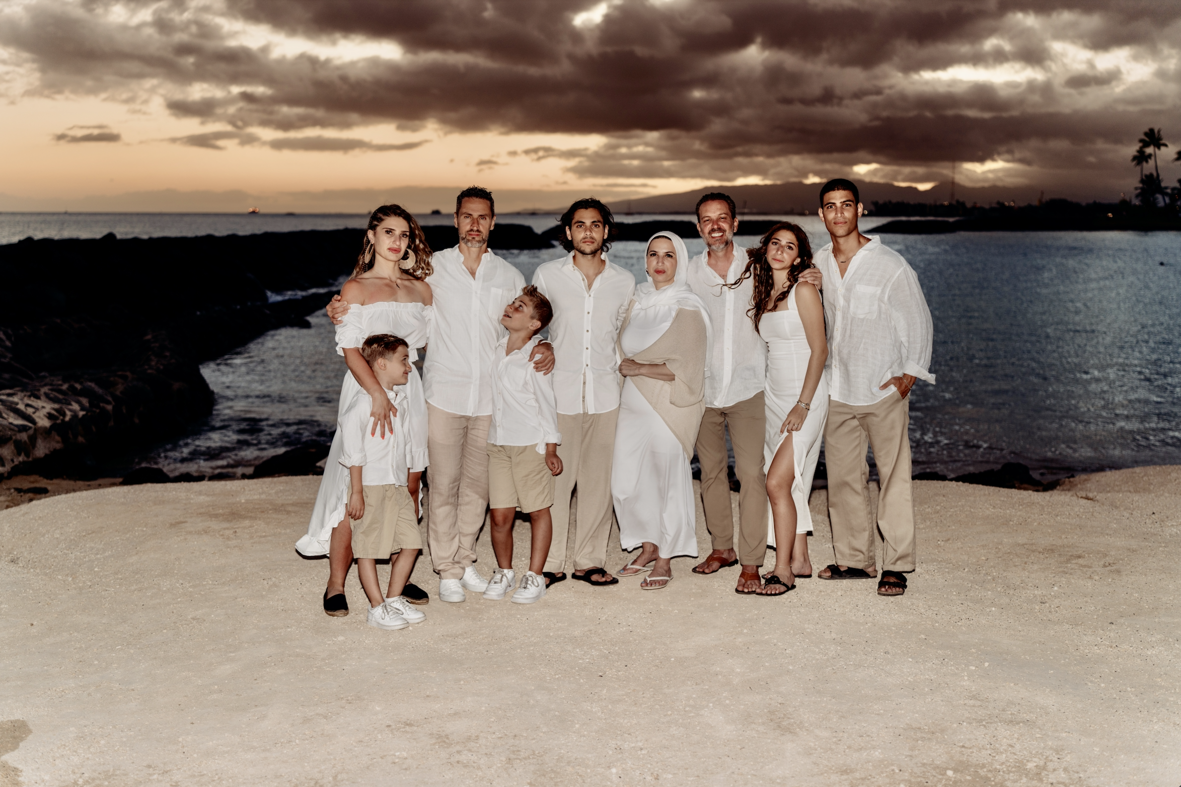 A family group dressed in white and beige clothing standing on a beach during sunset, with the ocean and cloudy sky in the background.