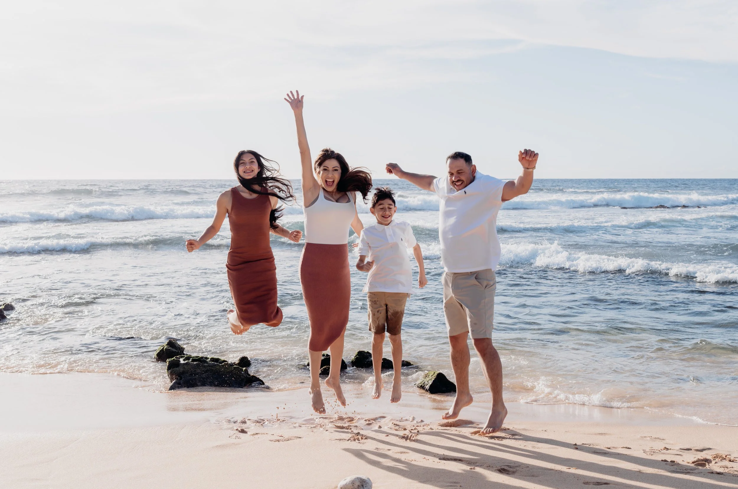 A family of four jumping on the beach with waves in the background, smiling and enjoying their time.