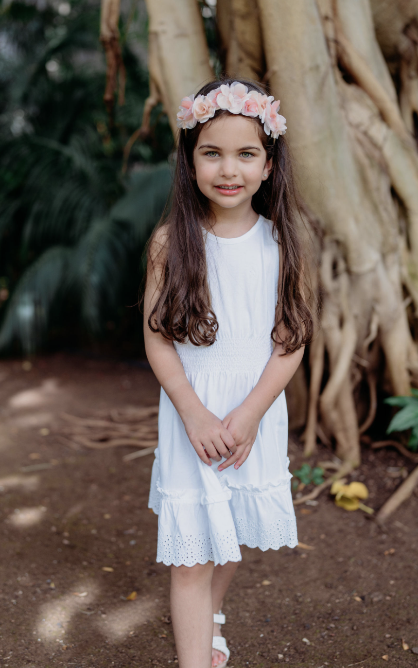 A young girl with long brown hair, wearing a white dress with eyelet details and a pink flower crown, stands outdoors in front of a large tree with exposed roots.