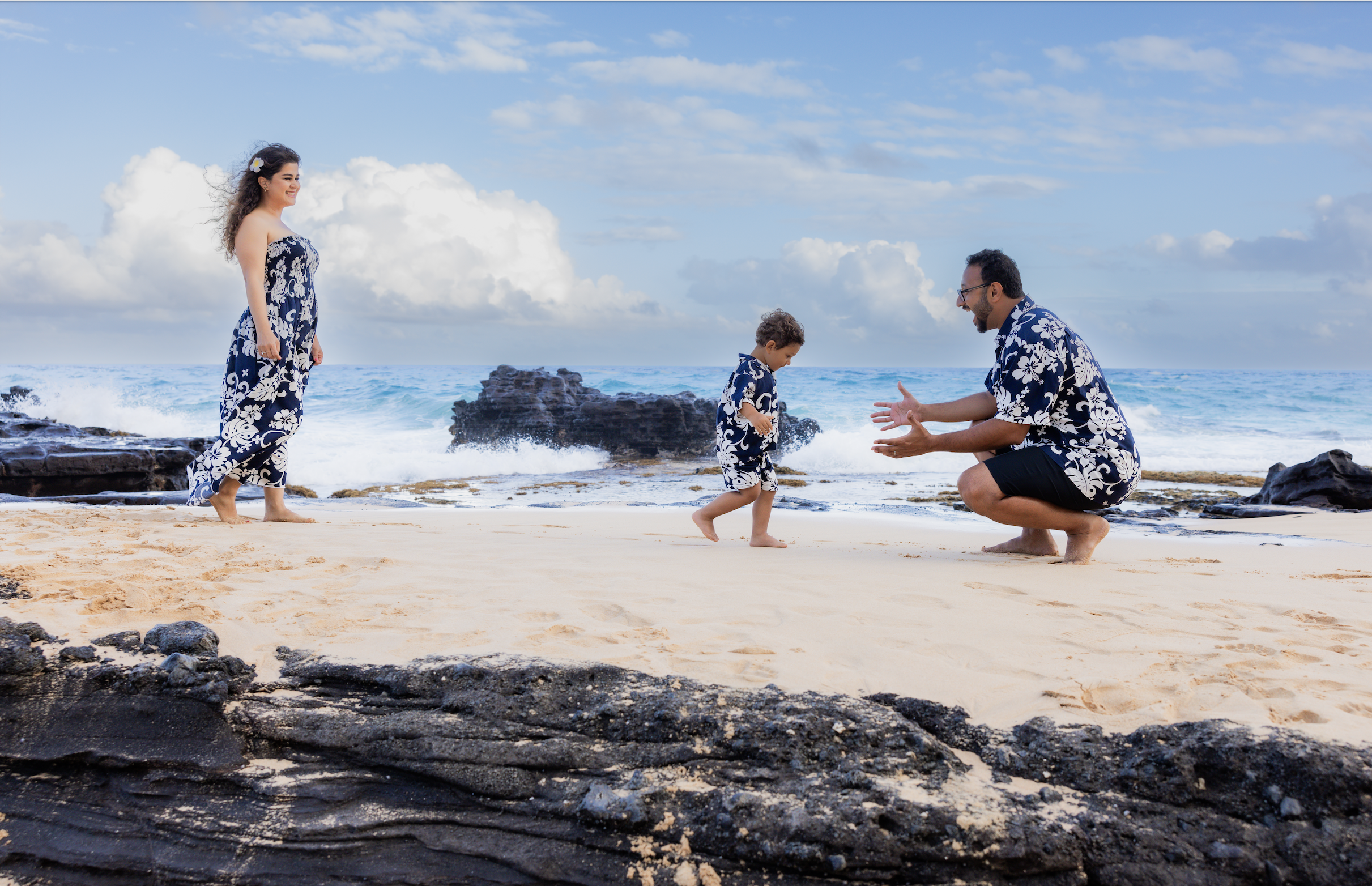 A family on a beach in matching blue and white Hawaiian shirts, with a woman standing on the left, a man squatting on the right with his arms extended, and a young boy in the center looking down, all smiling with the ocean and rocks in the background