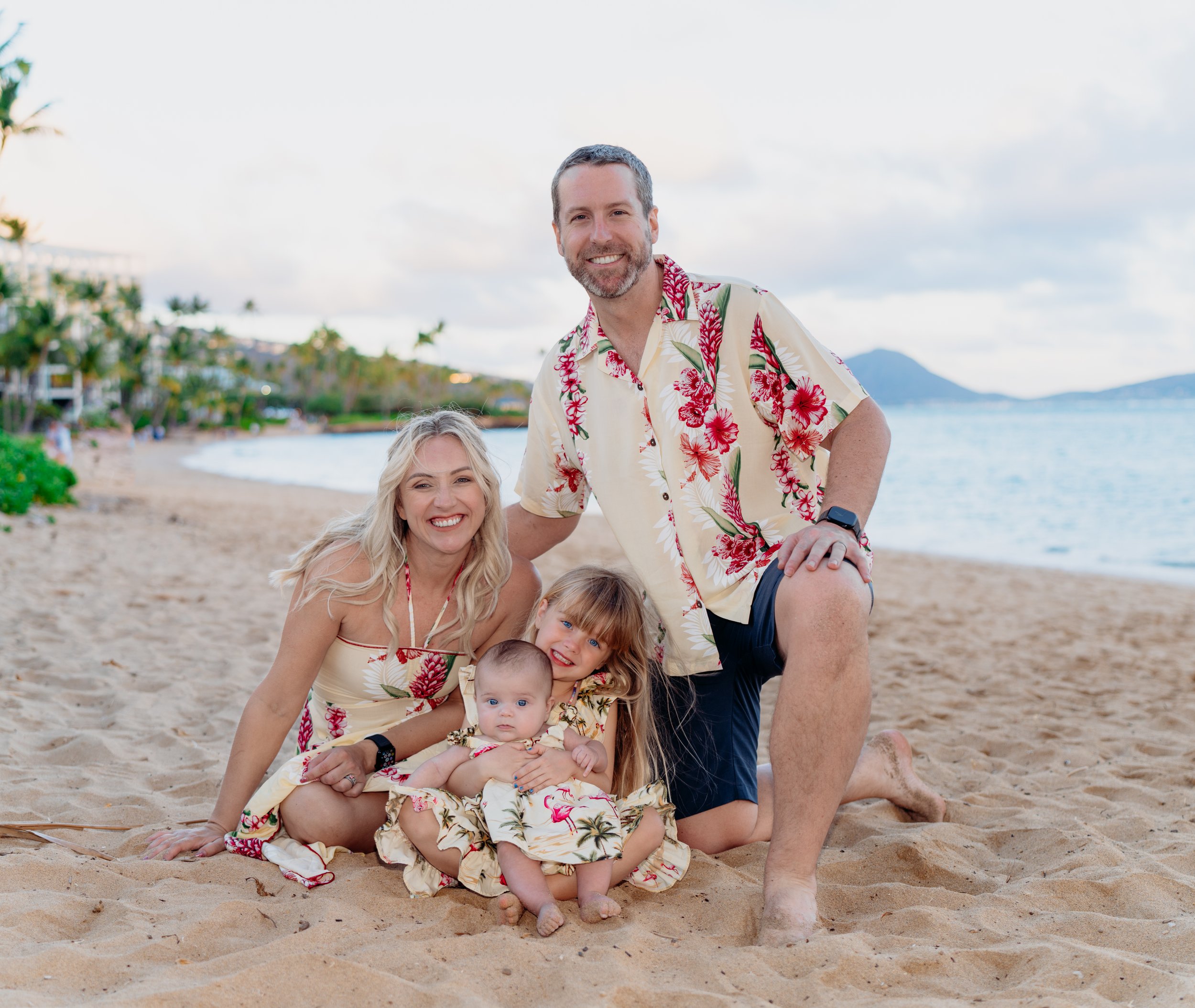 A family of four on a sandy beach, smiling, with the ocean and some islands in the background. They are wearing matching tropical print outfits.
