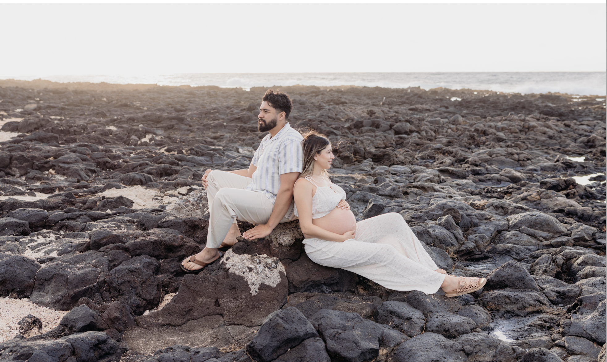 A pregnant woman and a man sit back-to-back on rocks by the beach during sunset, with the ocean in the background.