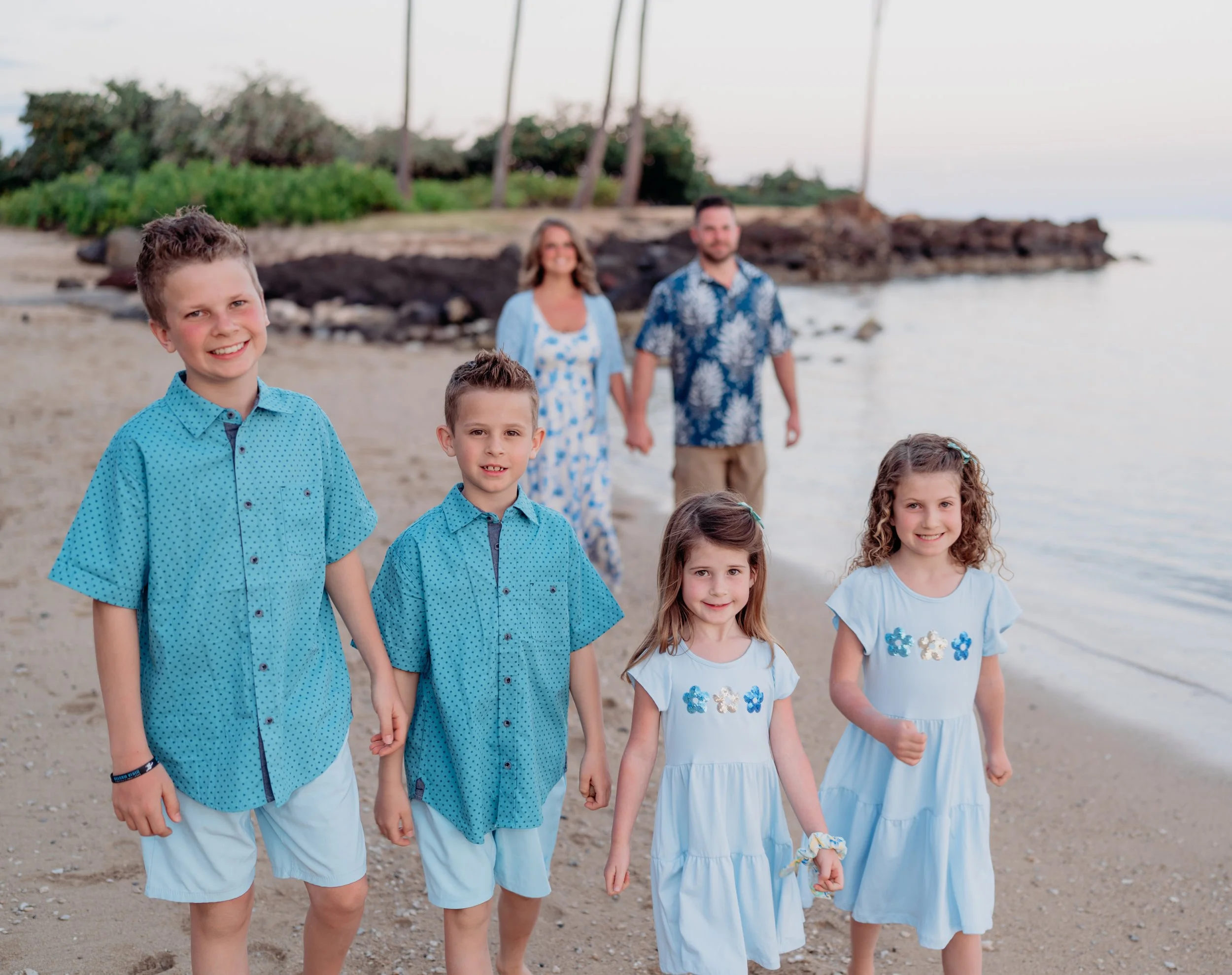 A family of seven with five children walking on a sandy beach near the water, holding hands, and smiling at the camera during sunset, with an ocean, trees, and a rocky shoreline in the background.