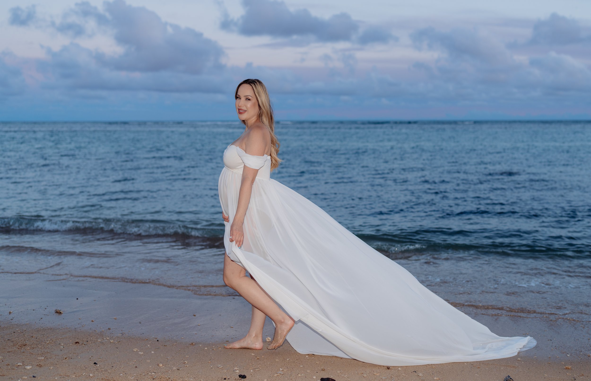 A woman in a white flowing dress stands barefoot on a sandy beach near the ocean with waves and cloudy sky in the background.