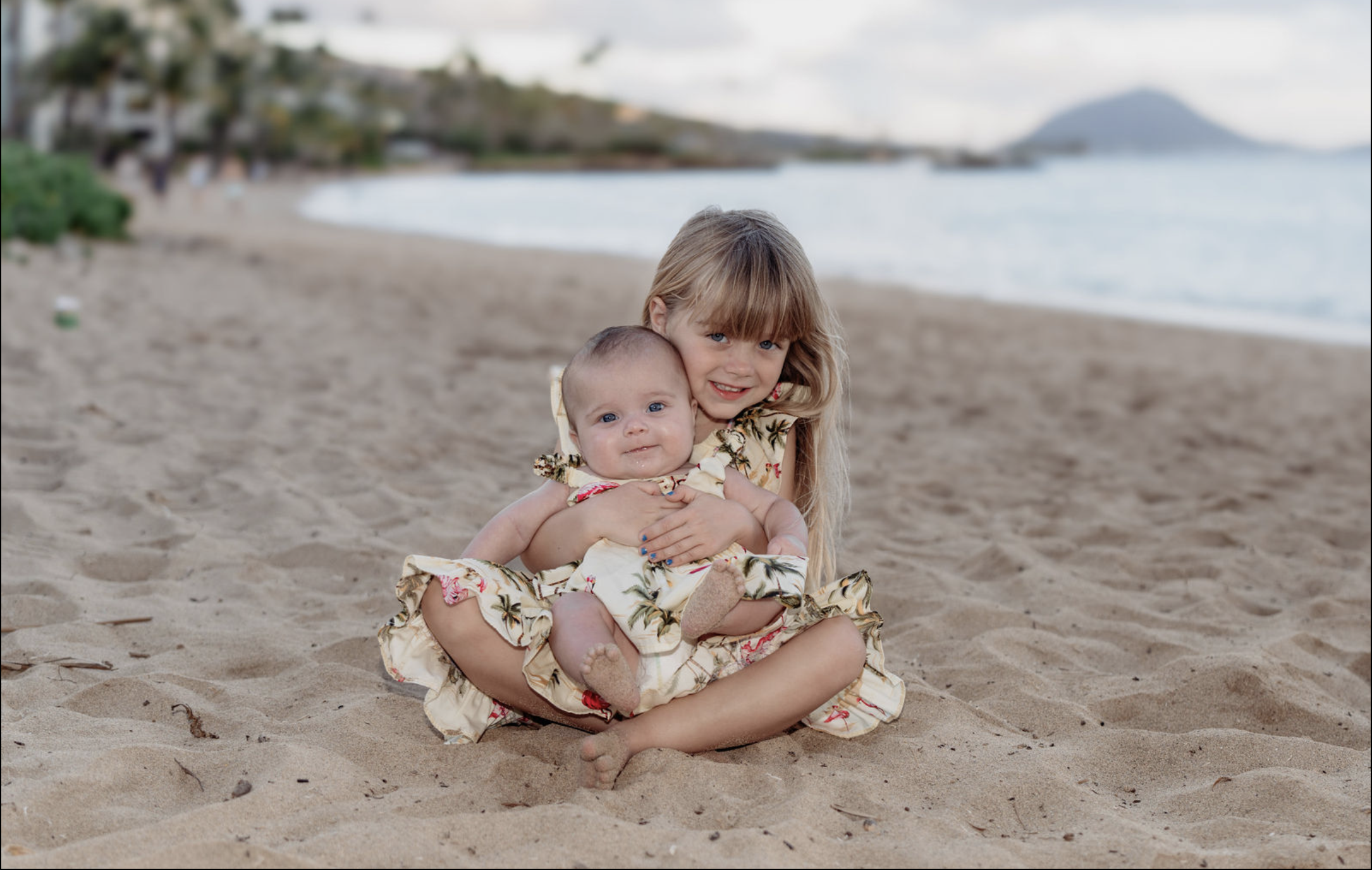 A young girl with blonde hair sitting on the sandy beach, holding a baby girl dressed in a floral dress, with the ocean and shoreline in the background.