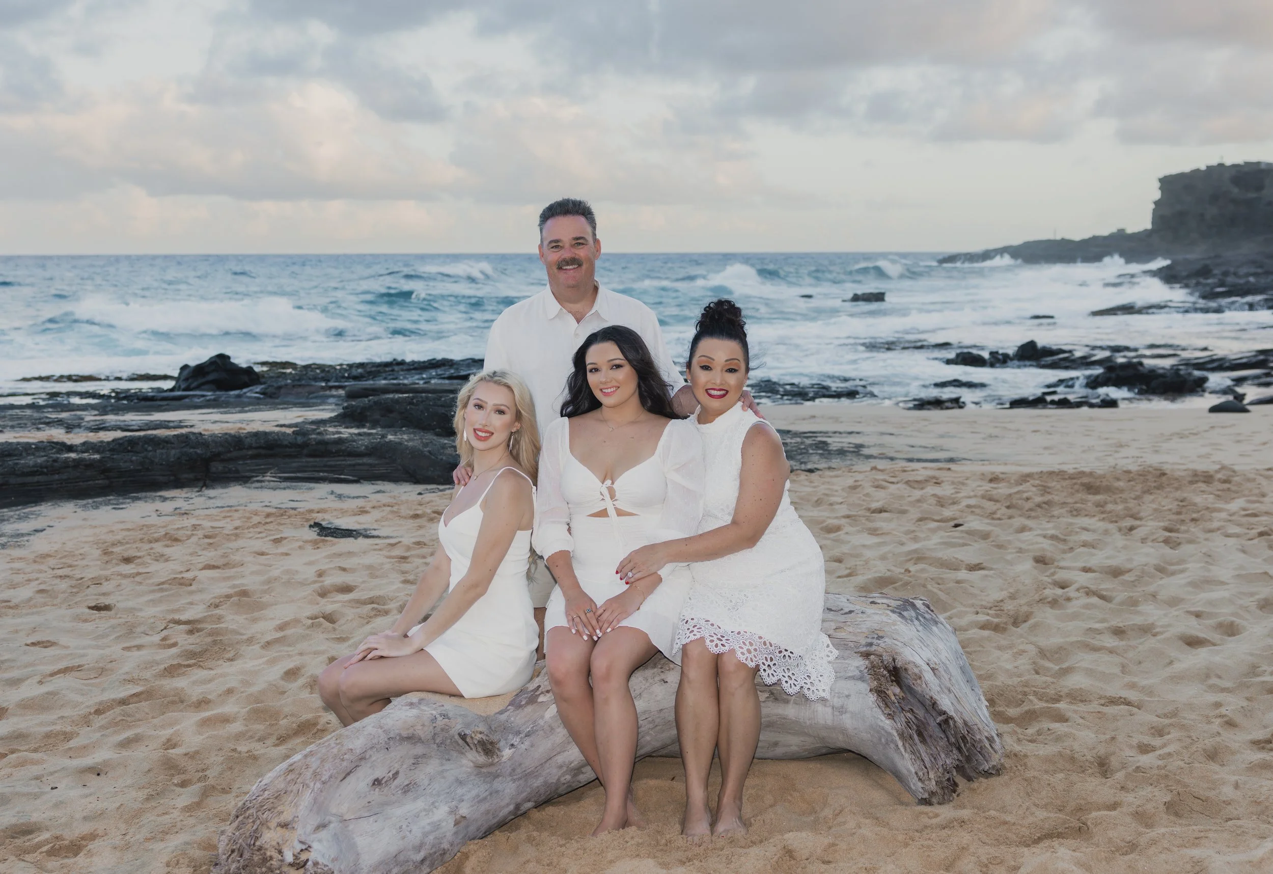 Group of four people sitting on a large piece of driftwood on a sandy beach with waves and rocky formations in the background.