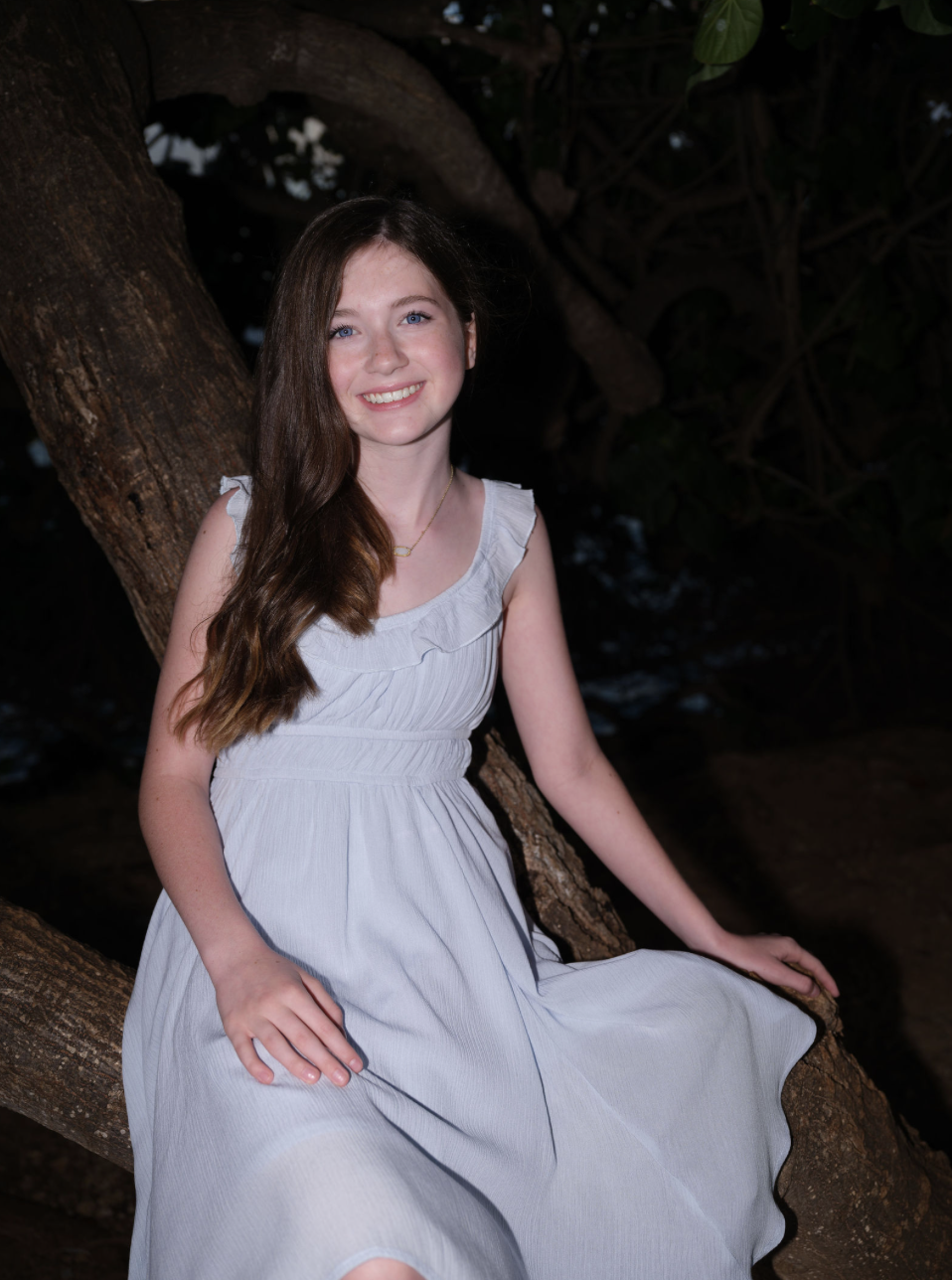 A young girl with long brown hair and blue eyes, smiling while sitting on a tree branch outdoors during dusk or evening, wearing a sleeveless white dress.