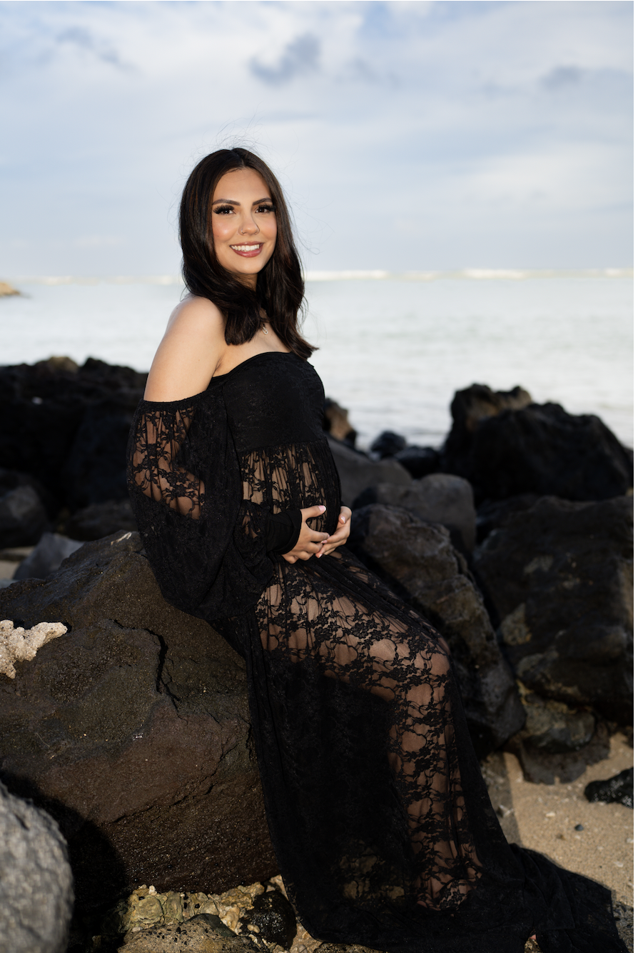 A pregnant woman sitting on rocks at the beach, wearing a black lace off-the-shoulder dress, smiling, with a cloudy sky and ocean in the background.