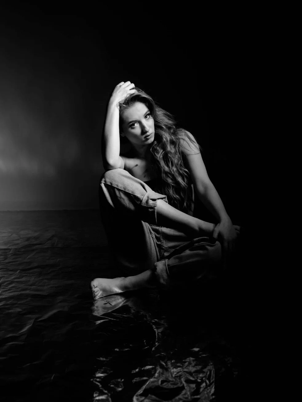Black and white photo of a woman sitting on a reflective surface, with one arm resting on her knee and her hand in her hair, looking at the camera with a serious expression.