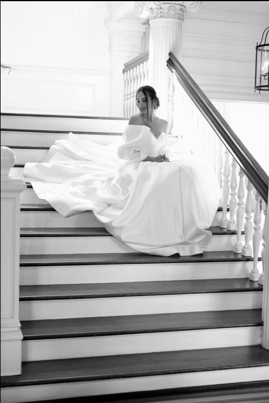 A woman in a white gown sitting on a staircase, with white railings and columns, in a bright, elegant interior setting.