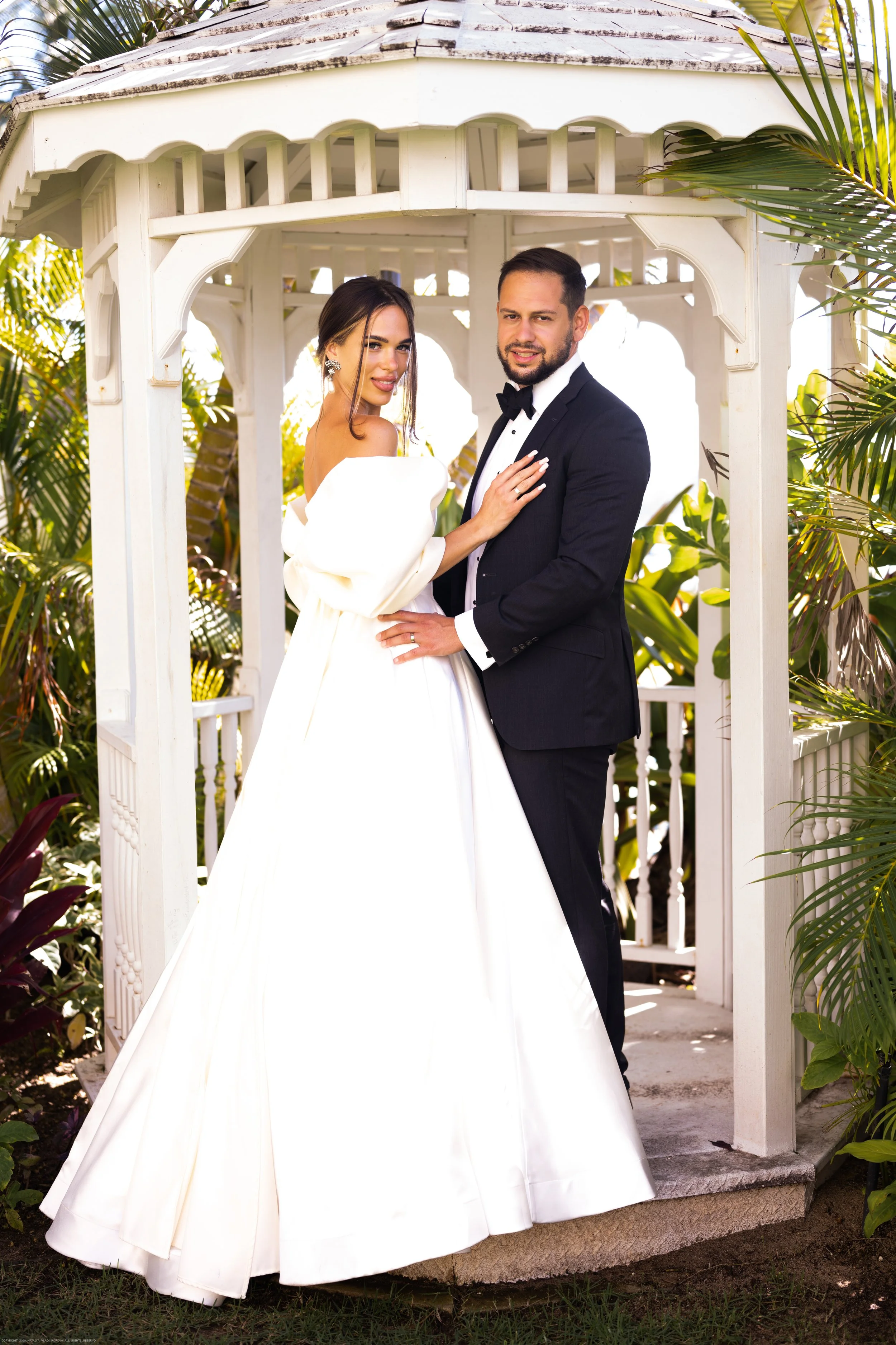 A bride and groom in wedding attire posing inside a white gazebo surrounded by tropical plants.