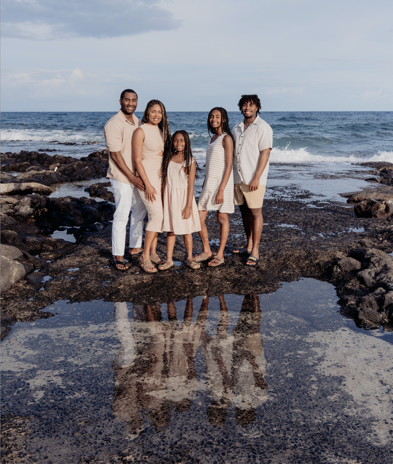 A family of five standing on rocks by the ocean at a beach, with waves and cloudy sky in the background. They are smiling and dressed in light, casual summer clothing.