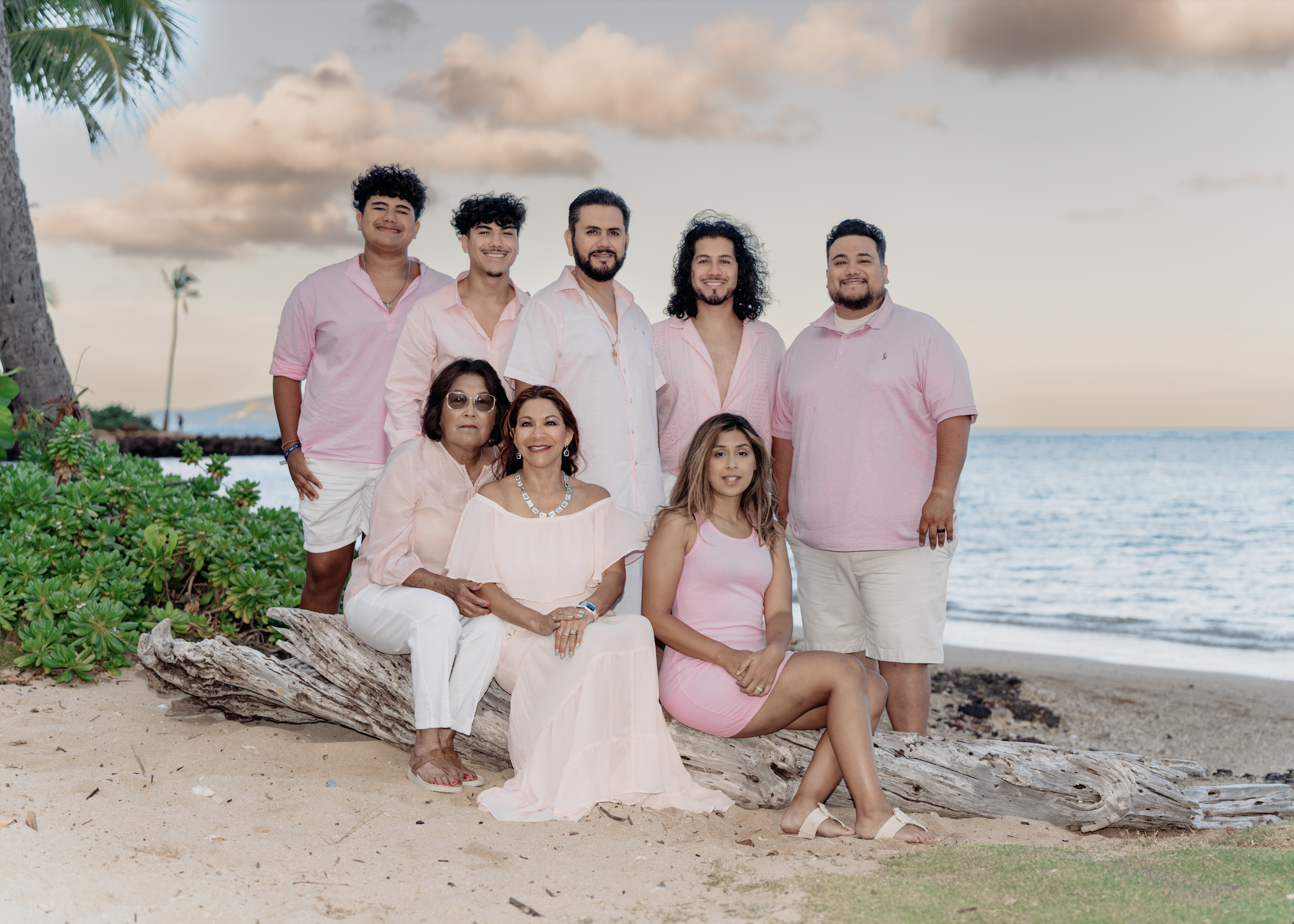 A group of nine people, dressed in pink and white, posing on a beach with a sunset in the background, surrounded by greenery and ocean.