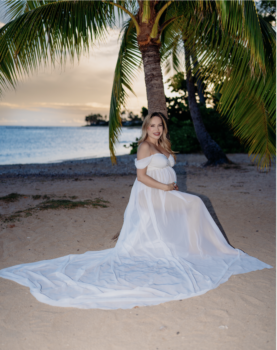 A woman in a white dress sitting on the sandy beach under a palm tree during sunset, with the ocean in the background.