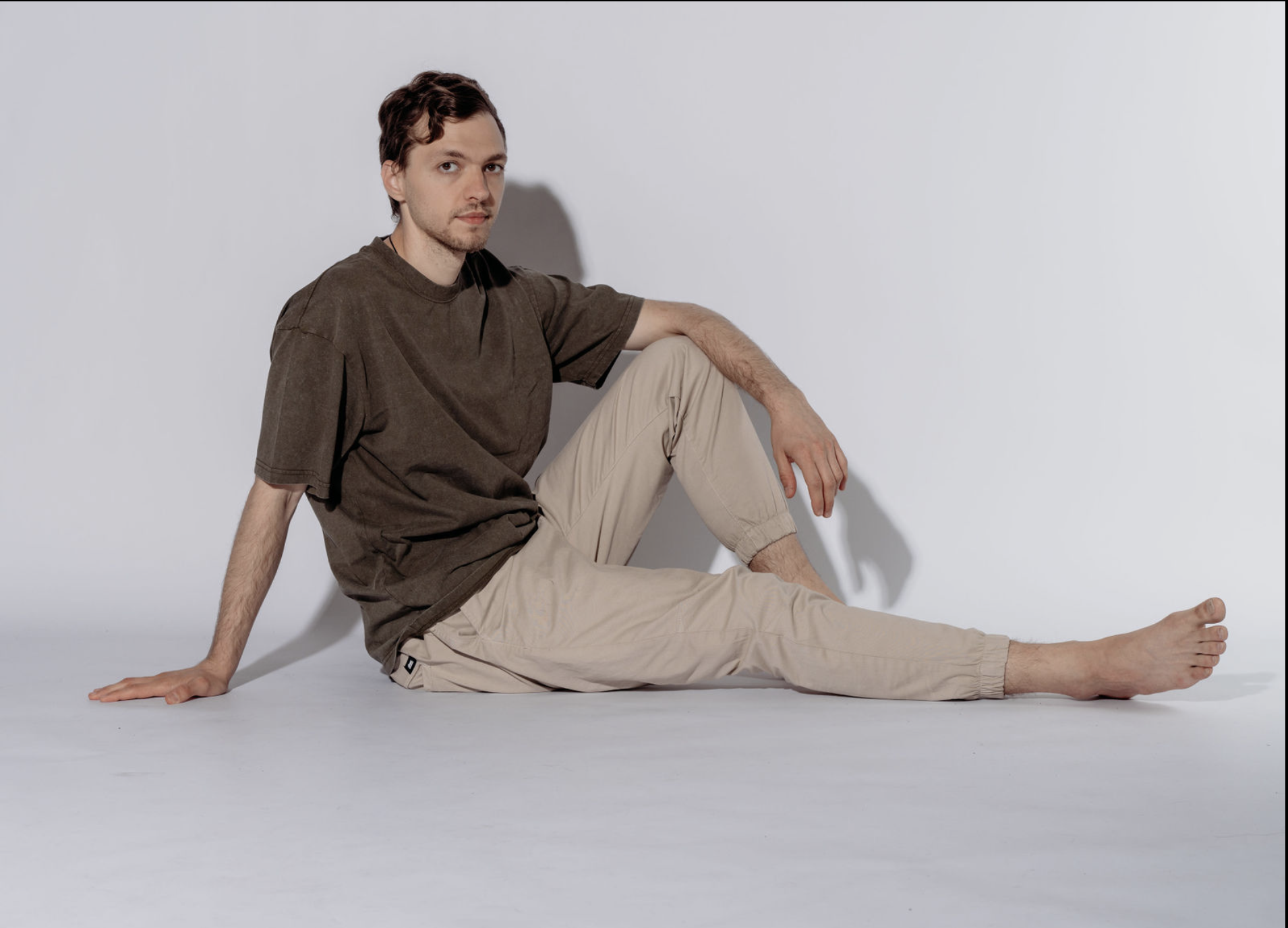 A young man with brown hair, sitting on the floor against a white wall, wearing a brown T-shirt and beige pants.