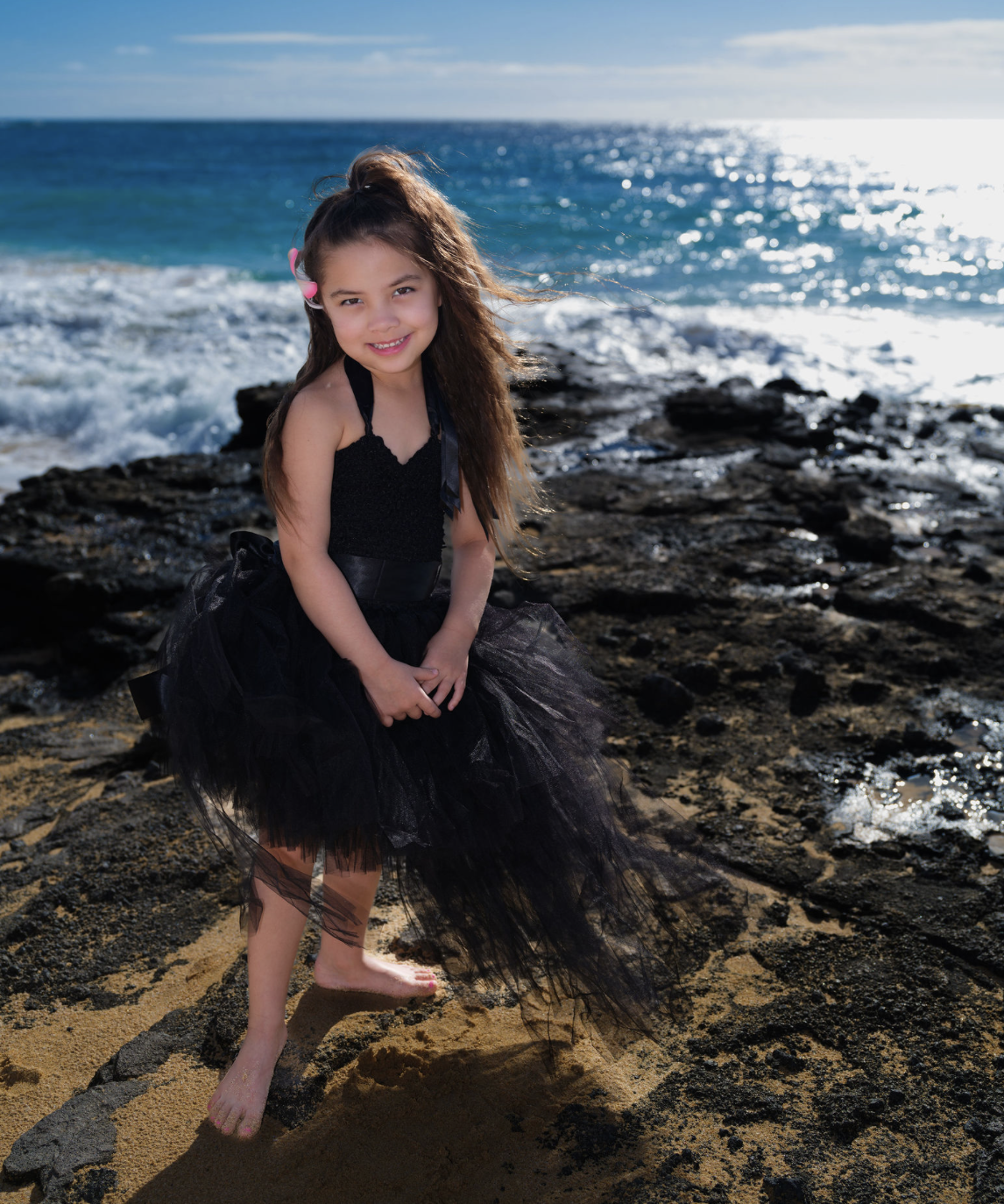 A young girl stands barefoot on a rocky beach wearing a black dress with a tulle skirt, with the ocean and a partly cloudy sky in the background.