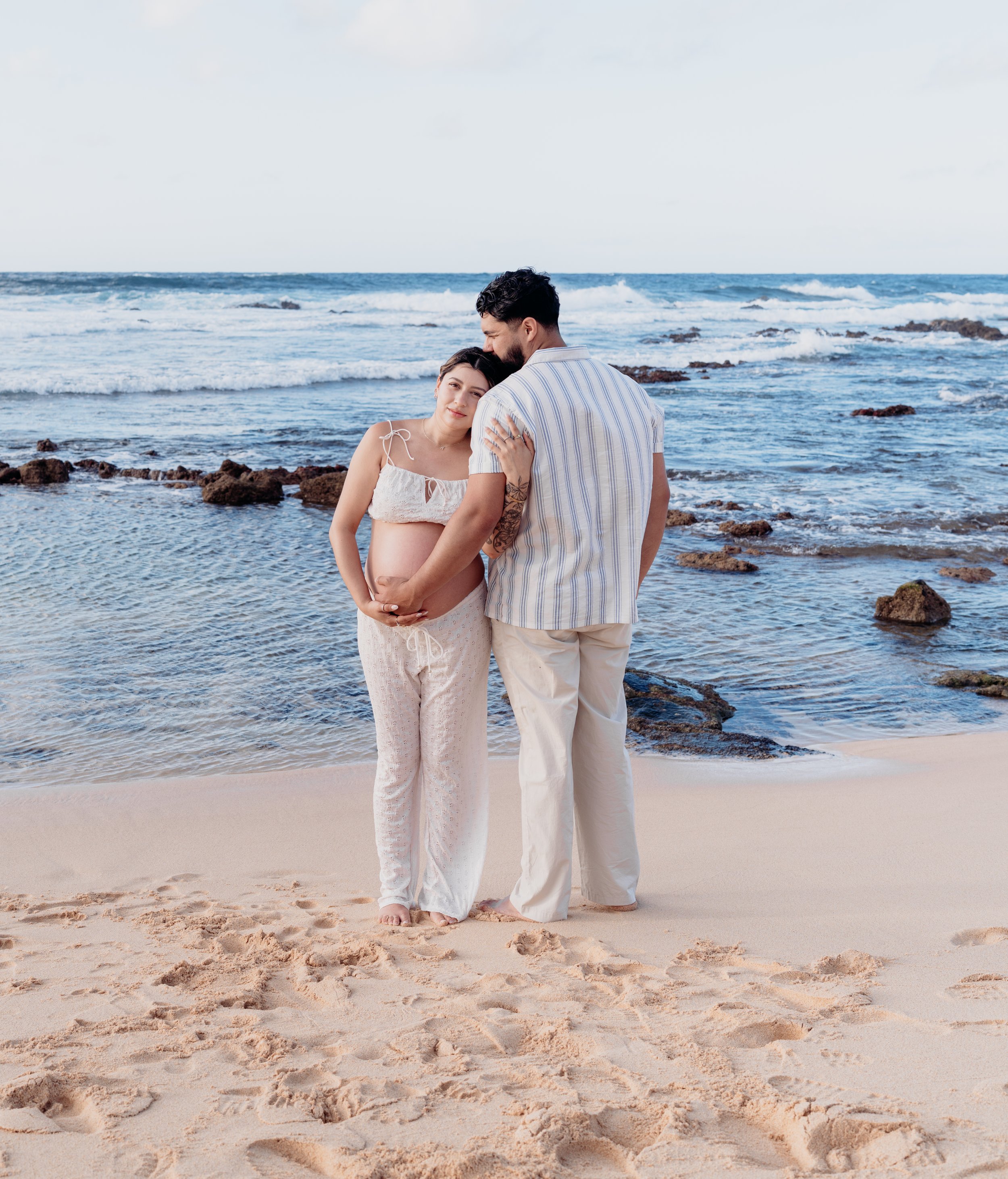 A pregnant woman and a man standing on a sandy beach with ocean waves and rocks, cuddling and looking at each other.