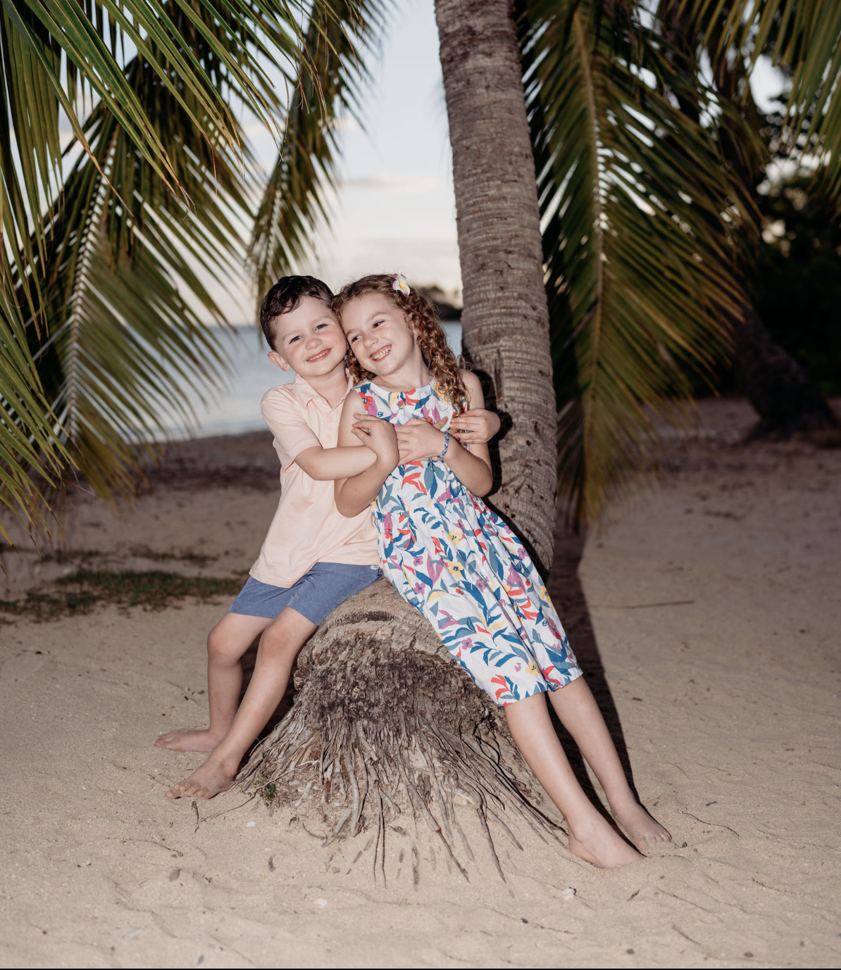 Two children, a boy and a girl, hugging and smiling on a tree stump at the beach with palm trees and the ocean in the background.