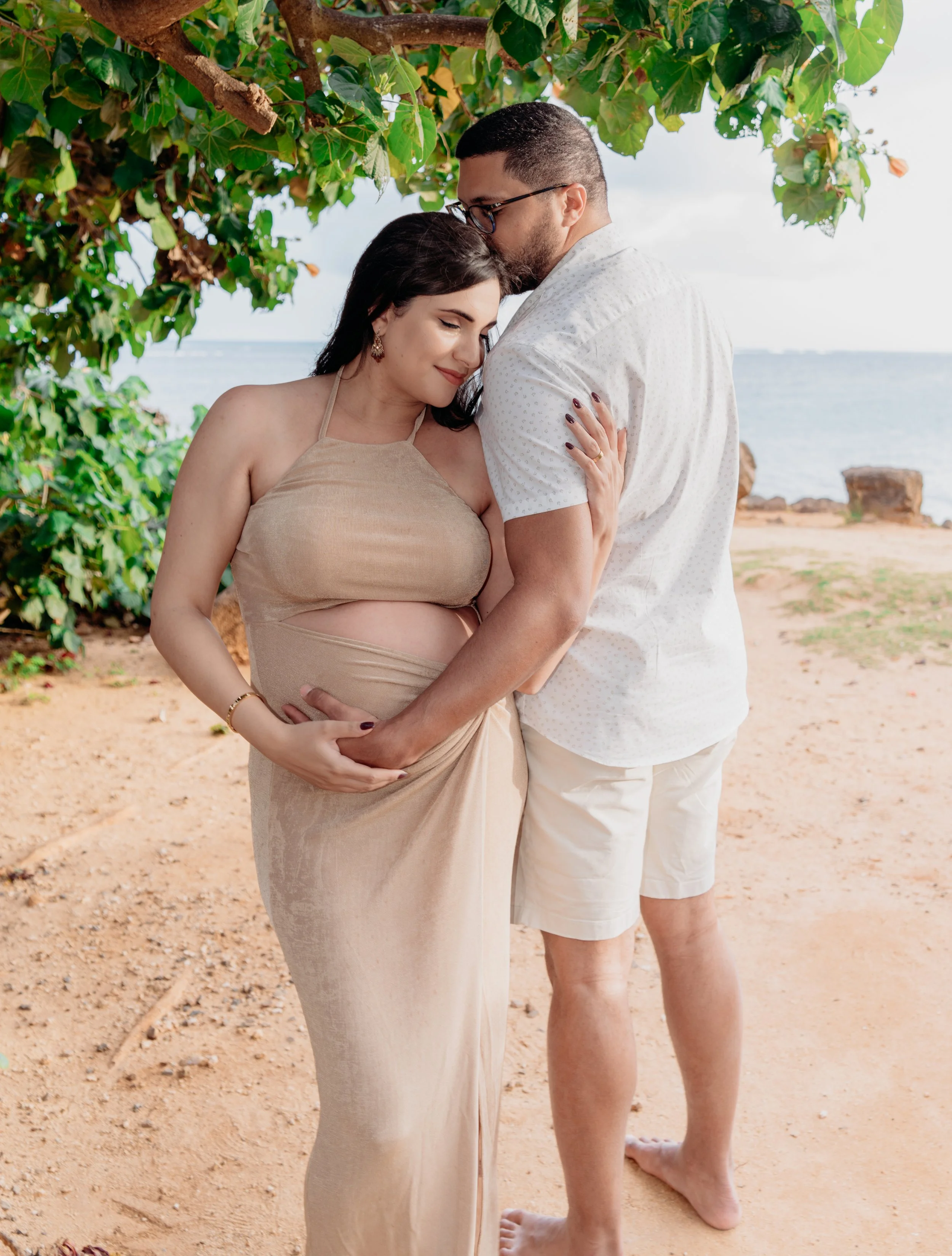 A pregnant woman and a man tenderly embracing on a beach, under a leafy tree, with the ocean in the background.