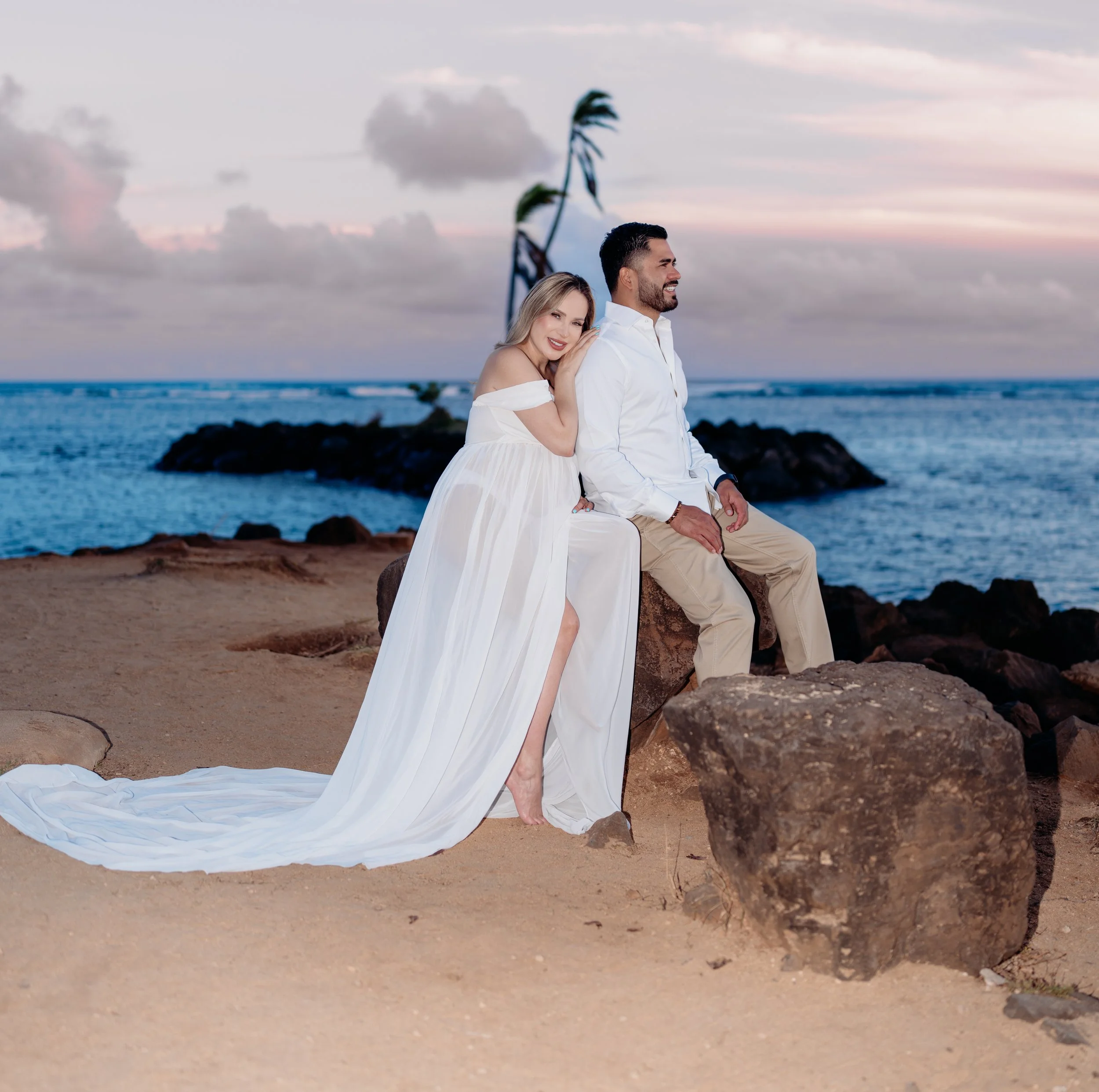 A couple on a beach during sunset, with the woman leaning on the man, both smiling, dressed in white and beige clothing, with a rock and a palm tree in the background.