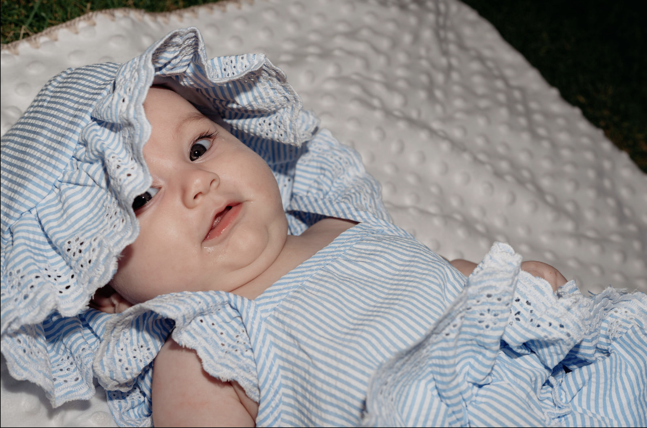 A baby lying on a textured white blanket, wearing a blue and white striped dress and matching bonnet.
