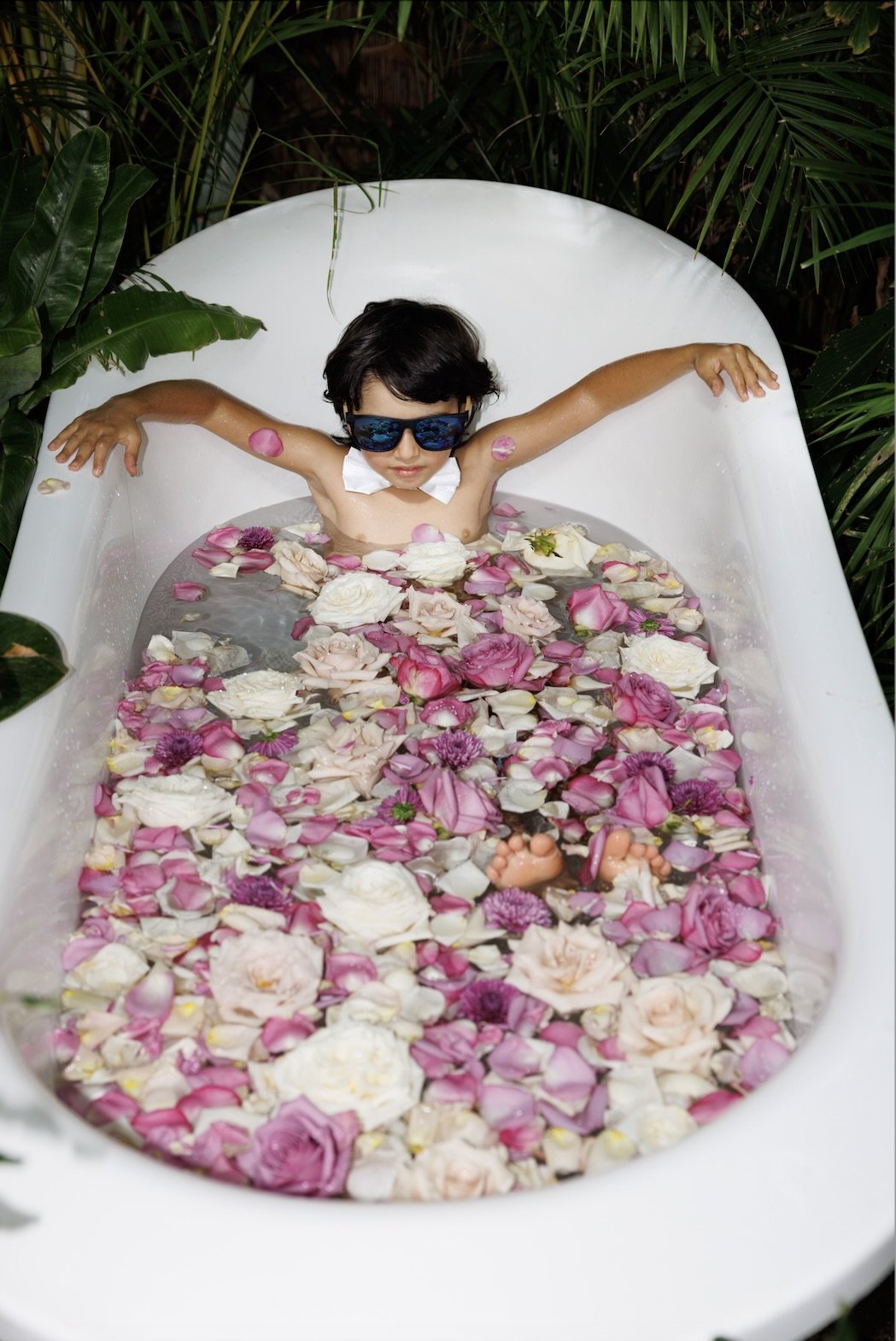Child wearing sunglasses relaxing in a bathtub filled with water and floating flower petals.