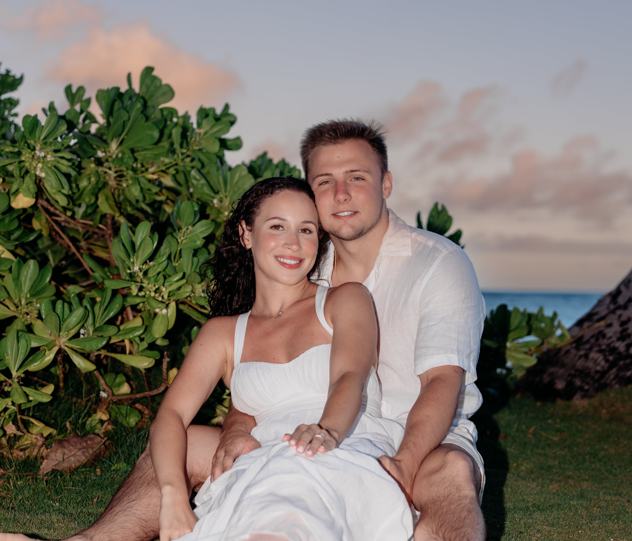 A smiling woman and a man sitting outdoors on grass near a bush and the ocean at sunset, wearing white clothing.
