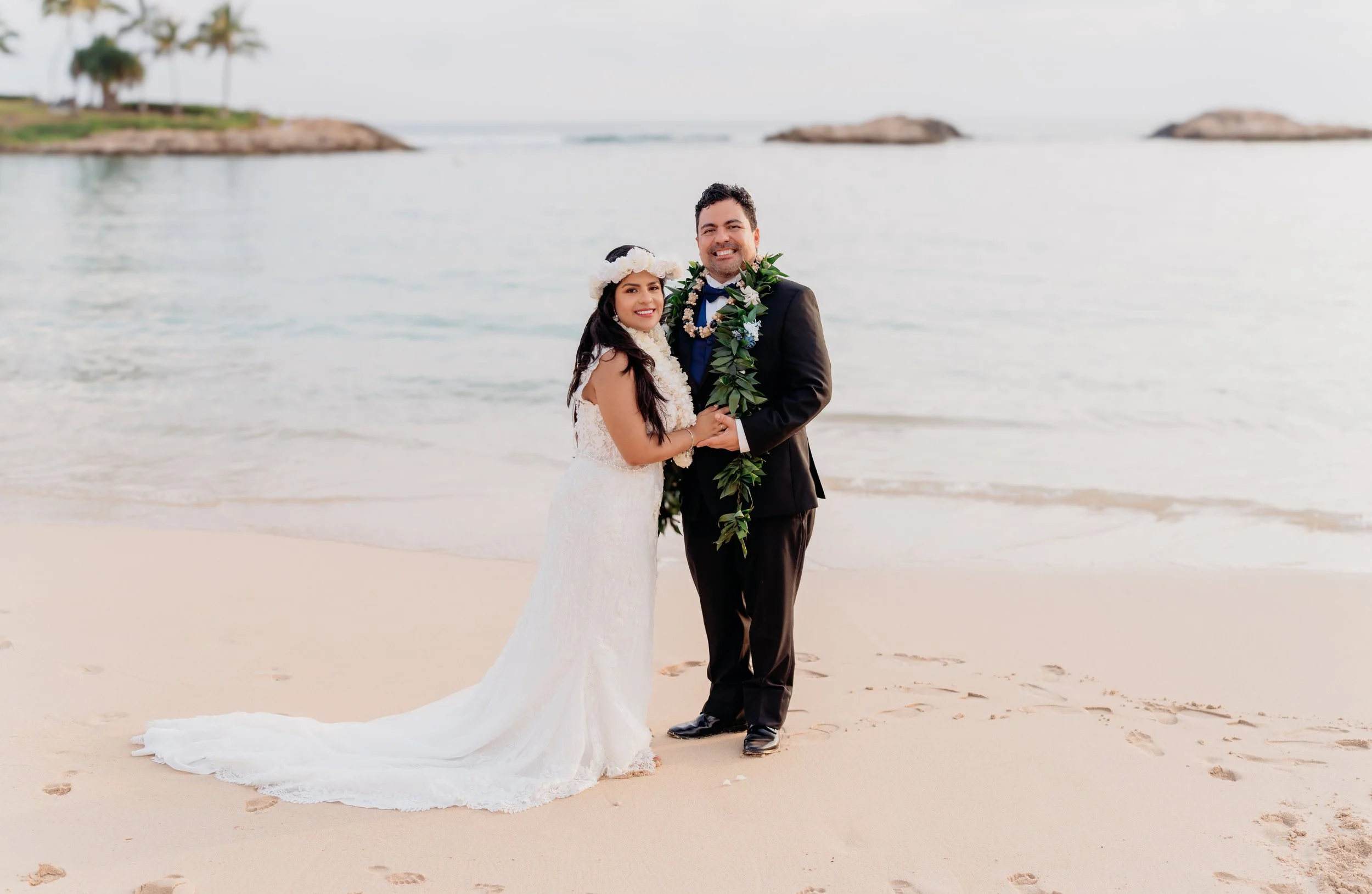 A newlywed couple in wedding attire holding hands on a sandy beach with ocean waves in the background.