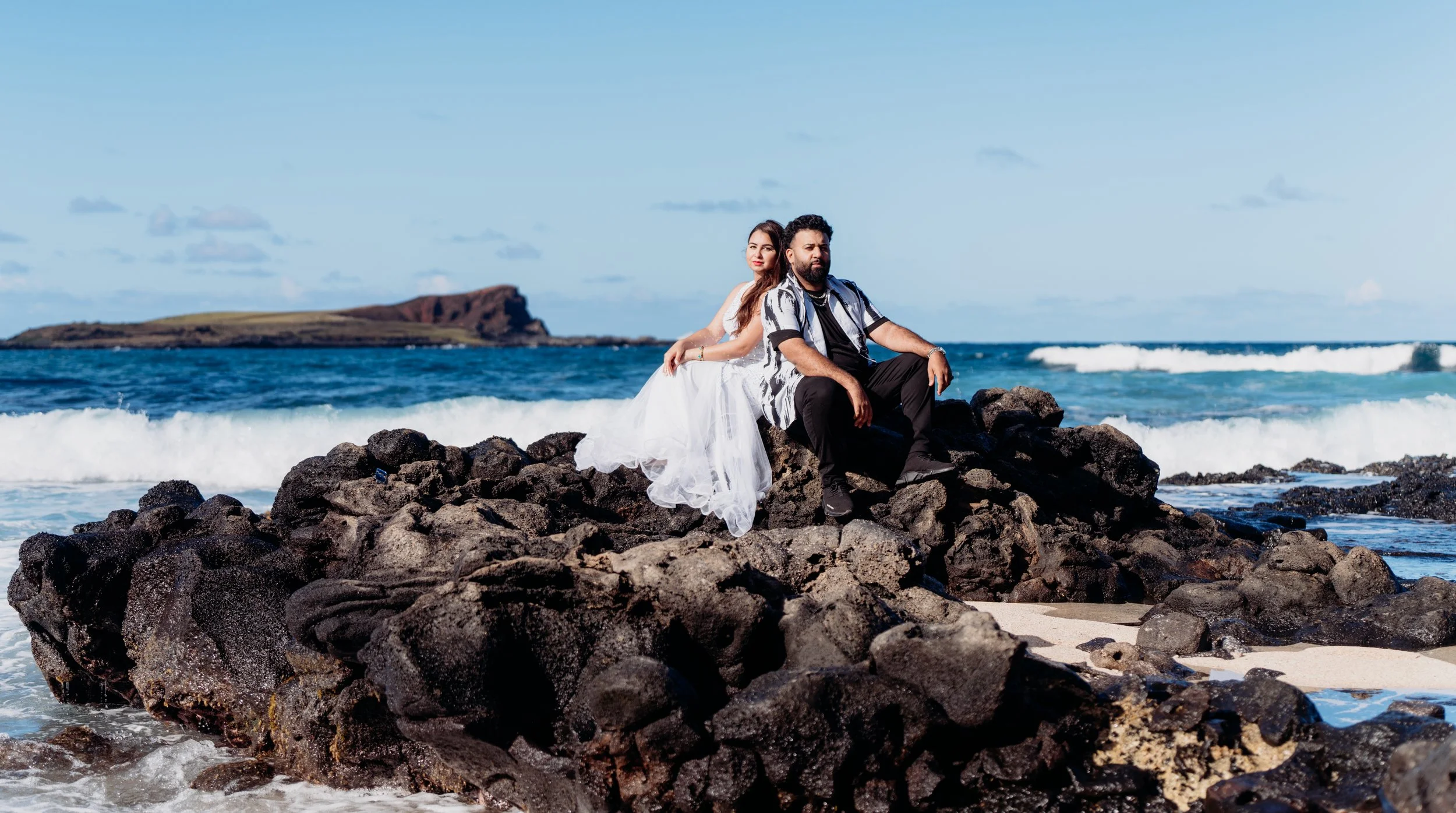 A couple sitting on black rocks at the beach, with the woman in a white dress and the man in a black and white shirt, with ocean waves and an island in the background.