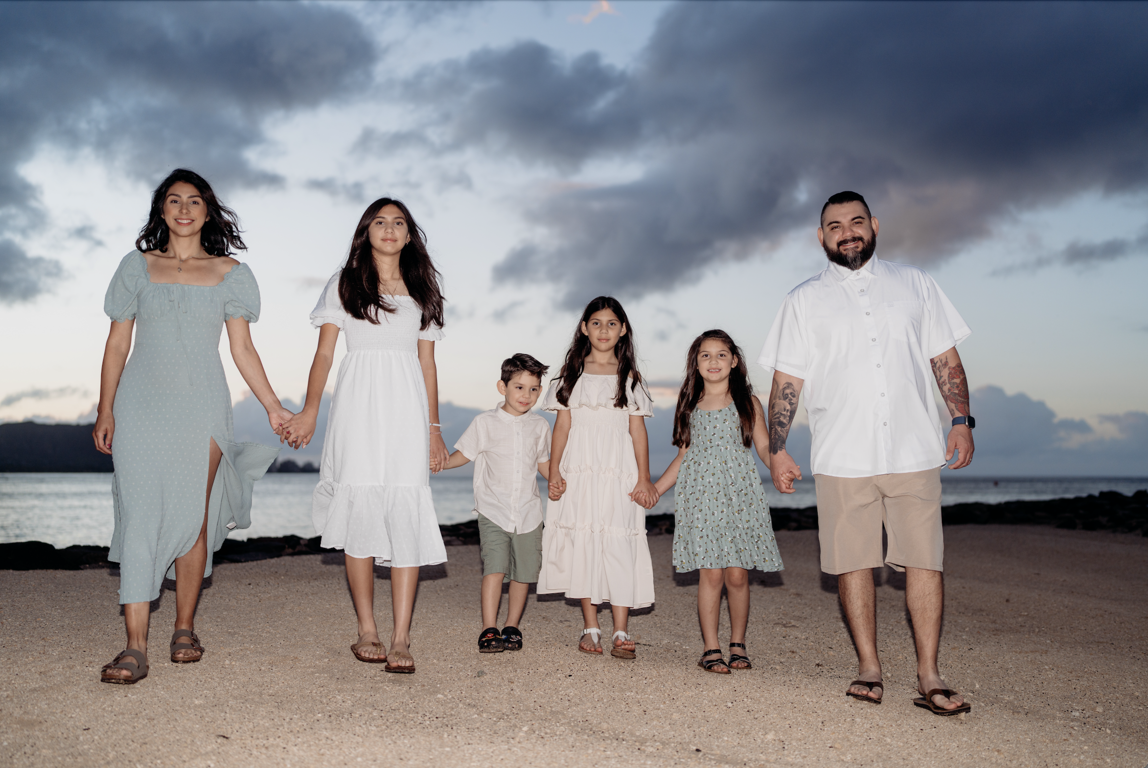 Family of seven walking on the beach at sunset, holding hands, with sky and ocean in background.