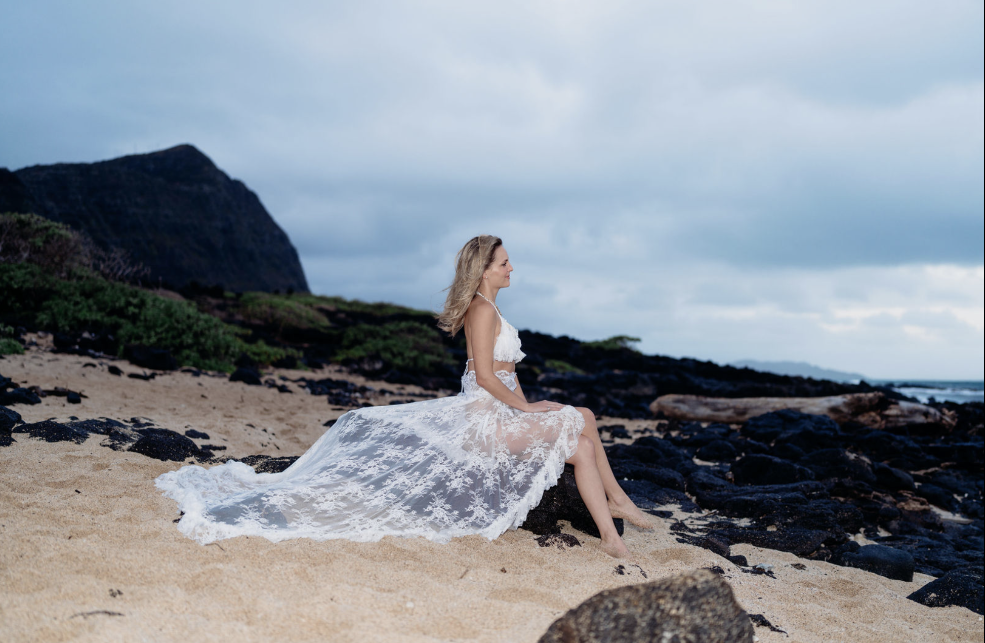 A woman in a white lace dress sitting on a rocky beach with sand, green shrubs, dark rocks, and cloudy sky in the background.